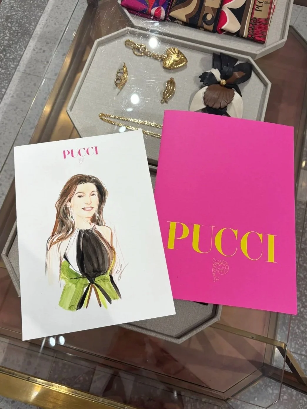 Jewelry display case with gold earrings, a necklace, and a bracelet. Two colorful Palm Angels gift boxes, and a Pink Pucci shopping bag. A fashion illustration of a woman with long hair, earrings, and a black and green outfit.