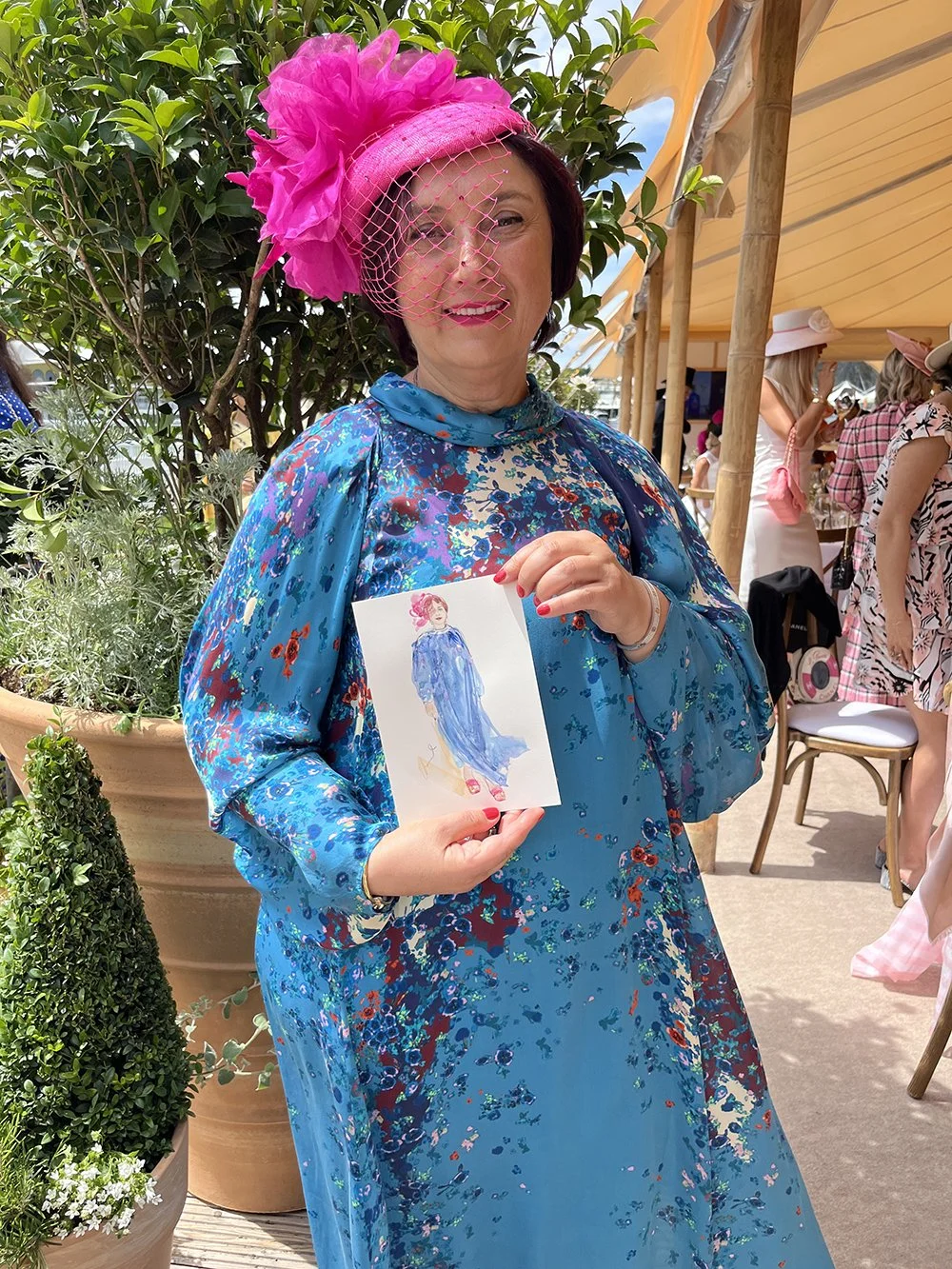 A woman in a vibrant blue floral dress and a large pink hat with a fascinator, holding a small illustration of a woman in a blue dress. She is outdoors at a gathering, with potted plants and other women dressed stylishly in the background.