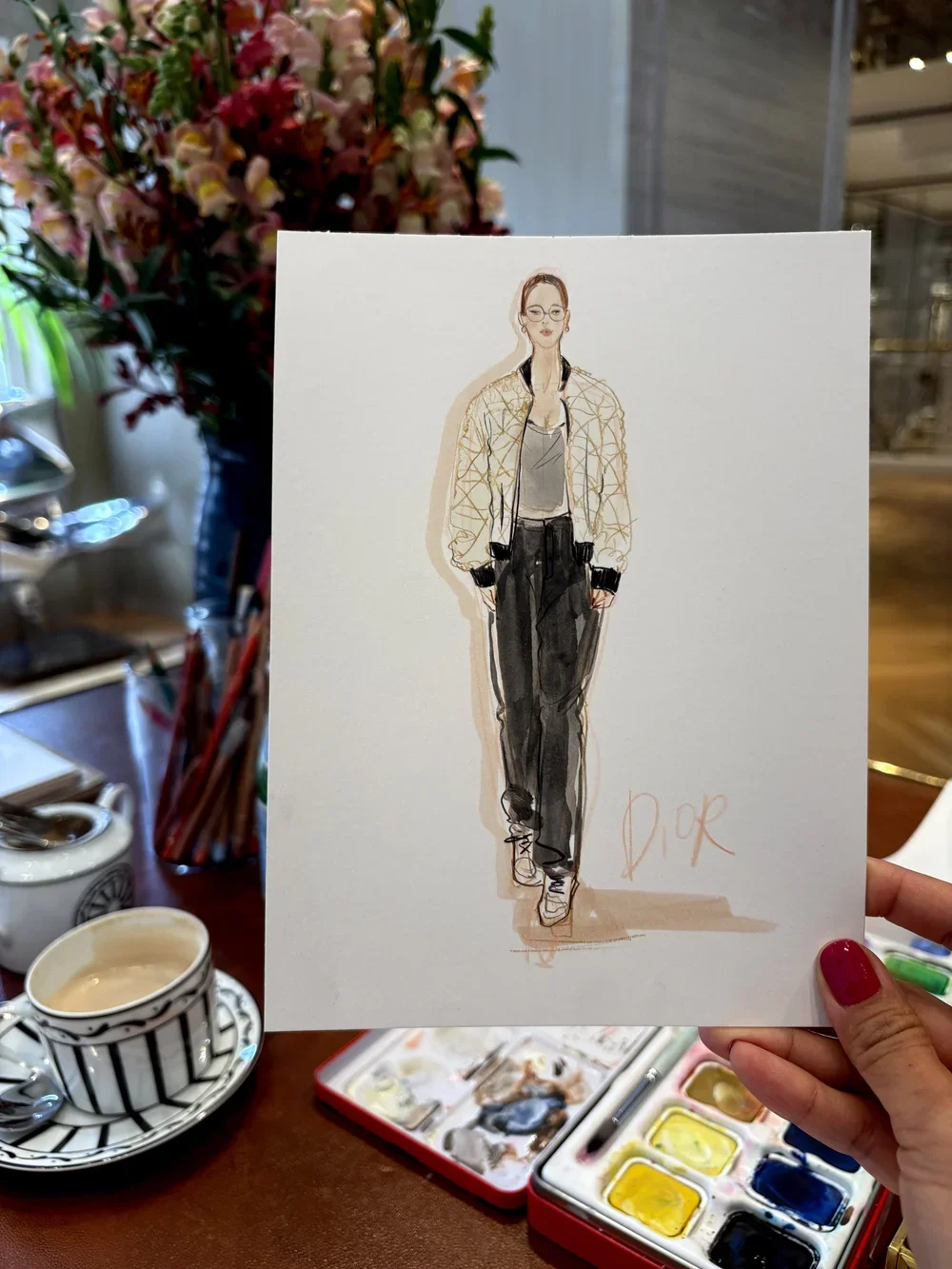 Hand holding a fashion illustration of a woman wearing glasses, a light jacket, gray top, and black pants, with the word 'Dior' written at the bottom. Background shows a floral arrangement, teacup, and watercolor paints.