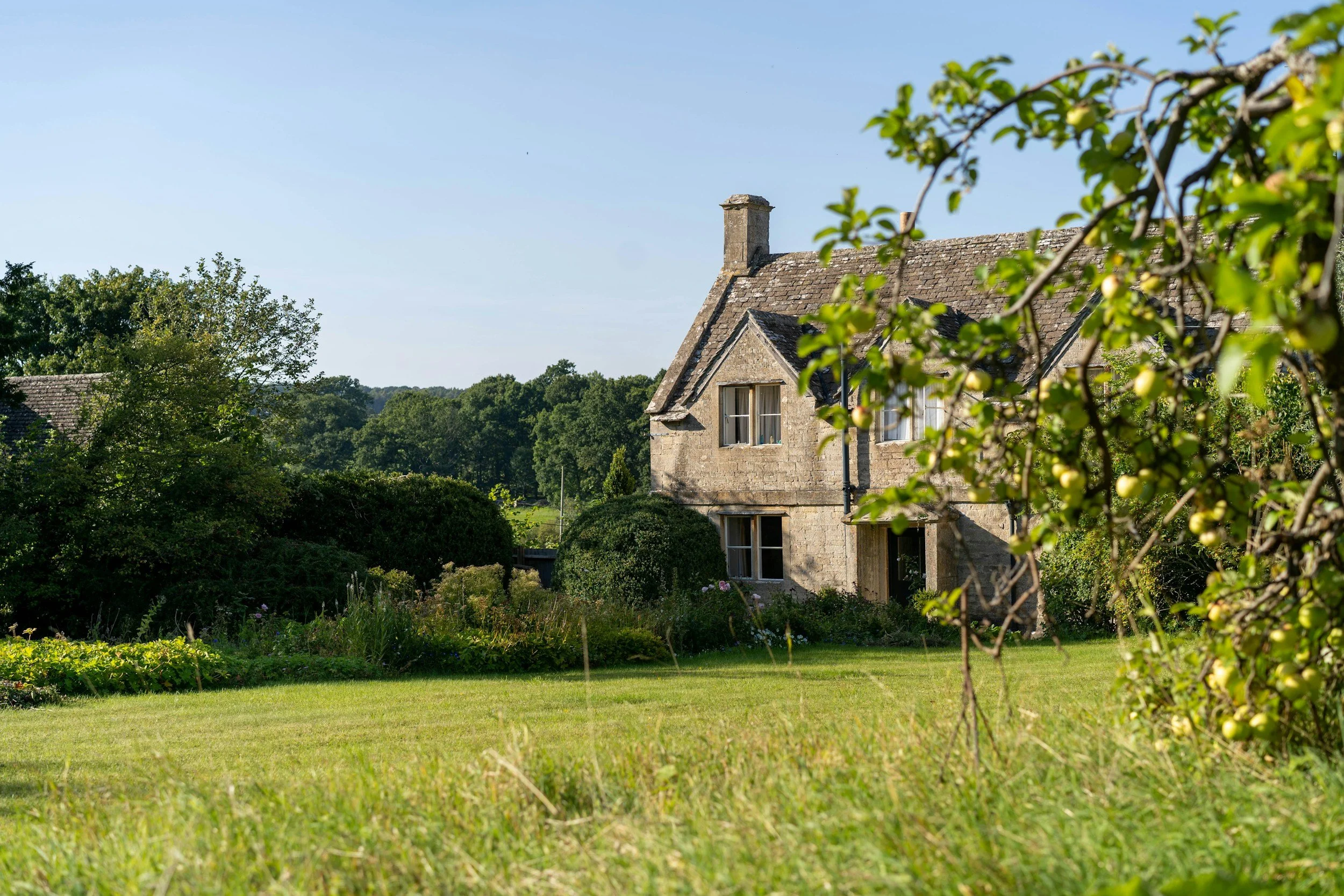 A stone house with a sloped roof and chimney, surrounded by green trees and a well-maintained lawn, with a clear blue sky above.