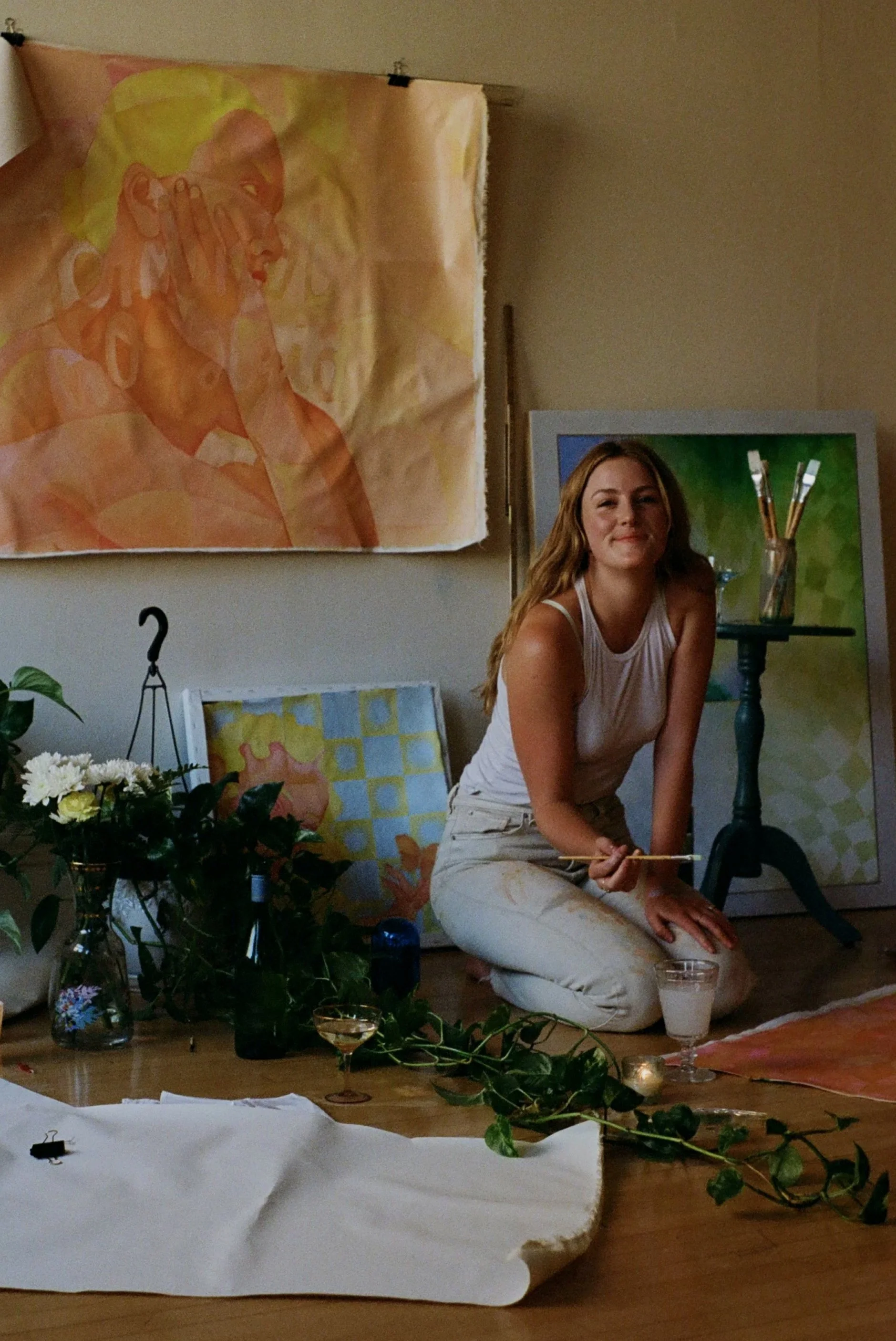 A woman kneeling on the floor in front of an art setup, with paintings and art supplies around her, holding a paintbrush and smiling at the camera.