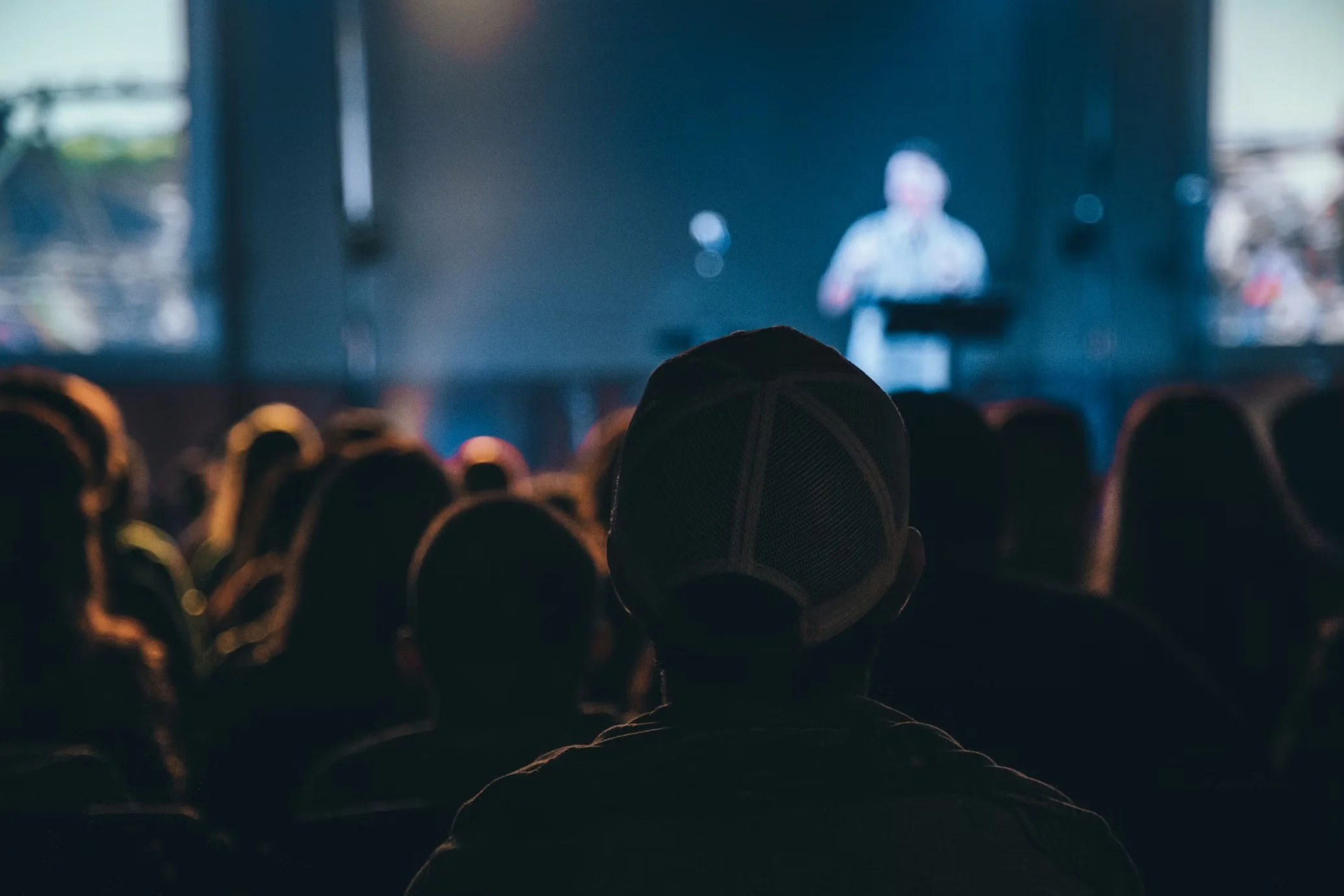 Audience watching a presentation or movie on a large screen in a dark theater or auditorium, with a person in the foreground wearing a baseball cap.