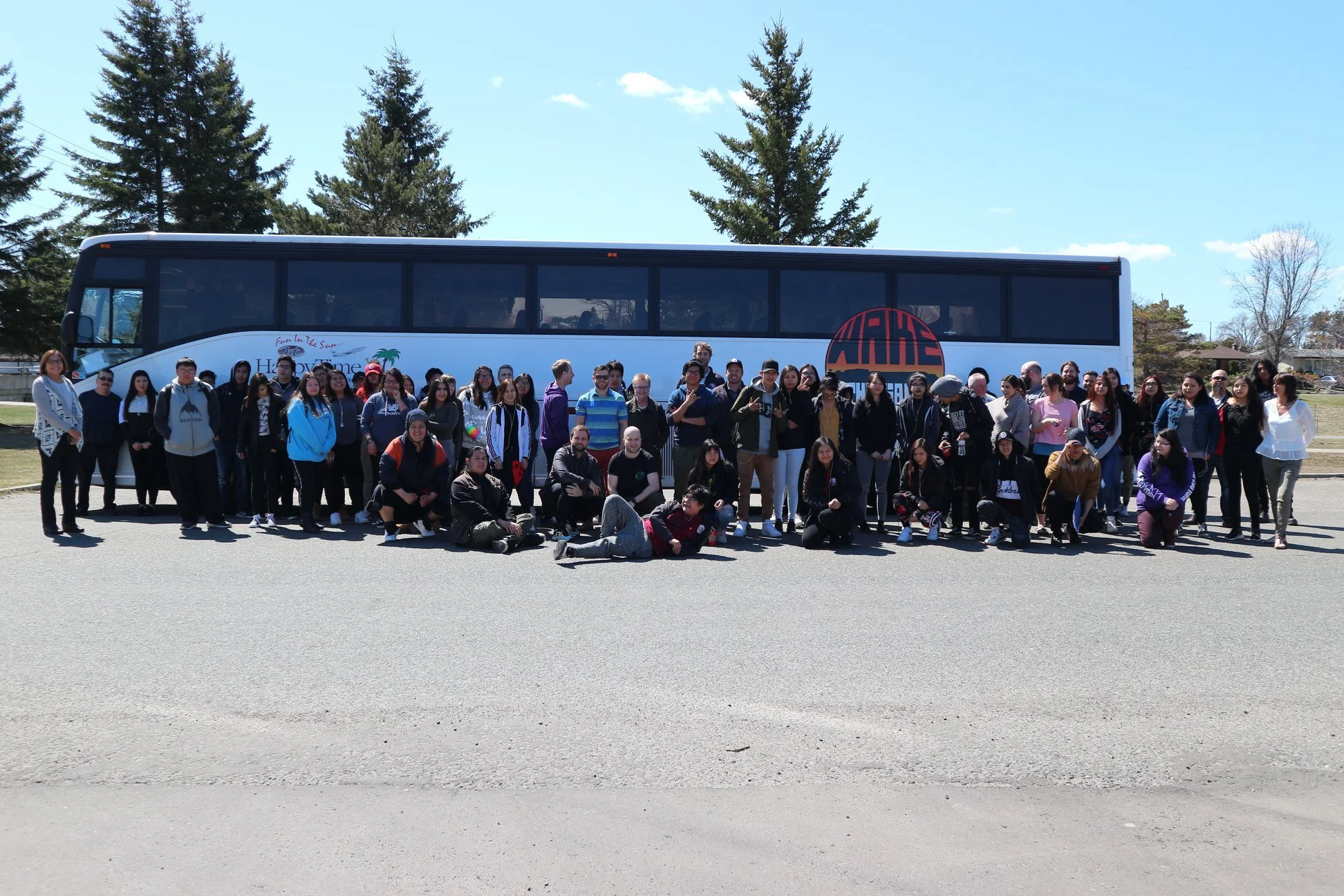 Group of people posing in front of a large tour bus with trees in the background on a sunny day.