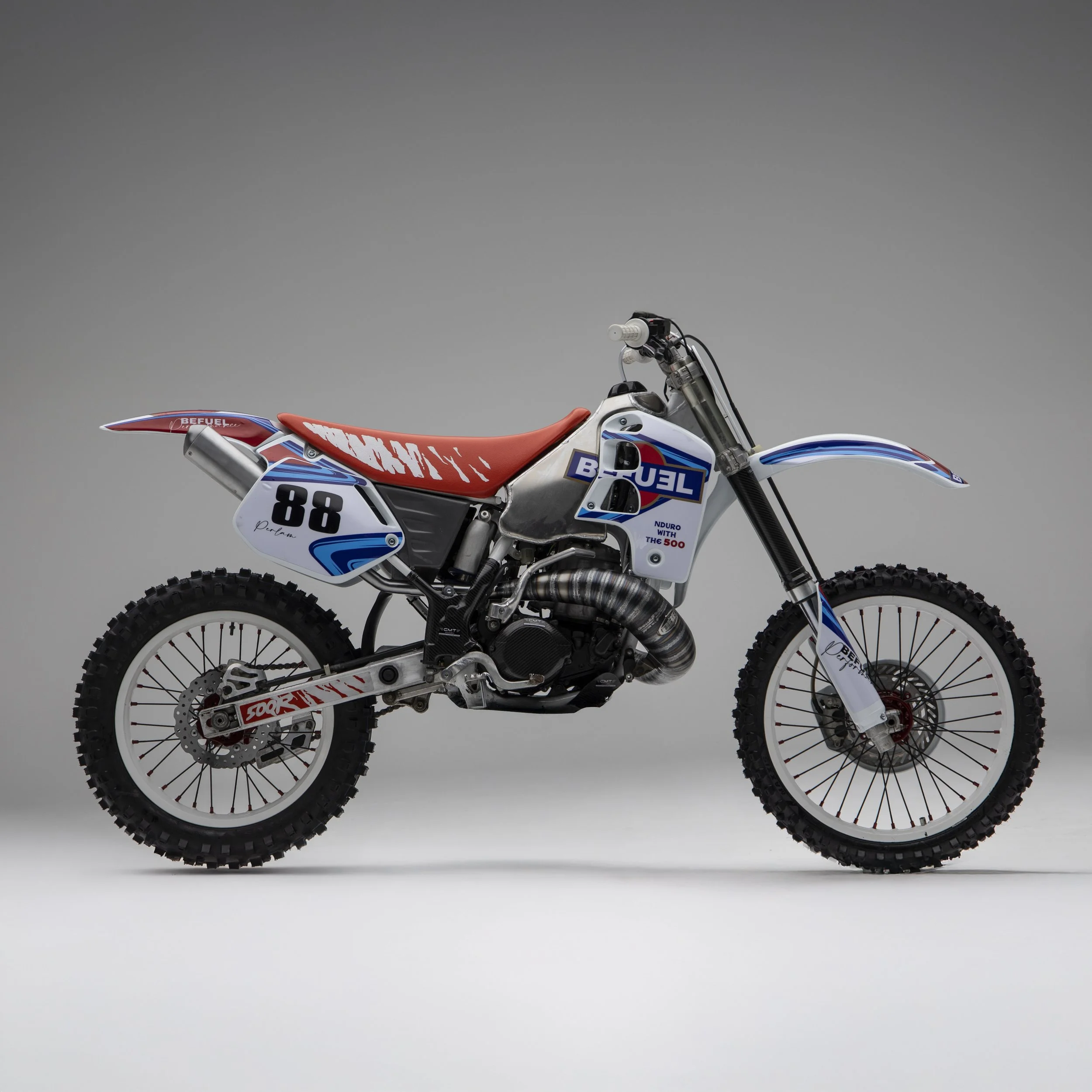 Motocross con telaio bianco, sella arancione, numeri 88, e decorazioni blu e rosse, su sfondo grigio.
