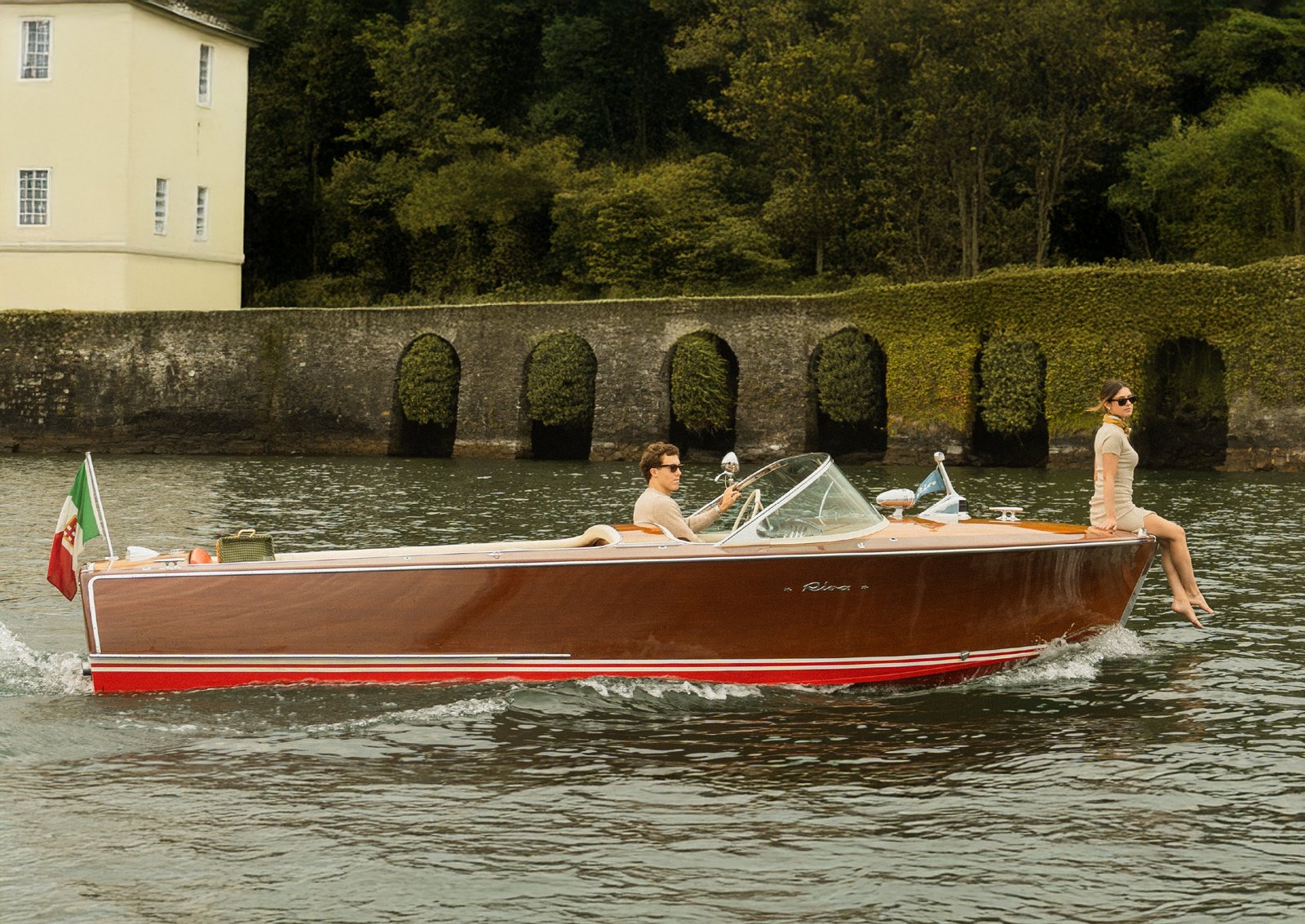 Una barca a motore con due persone a bordo, una donna seduta all'estremità della barca con le gambe penzoloni sull'acqua e un uomo seduto dietro il timone, in un corso d'acqua vicino a una sponda con un muro di pietra e un edificio beige, con un bosco sullo sfondo.
