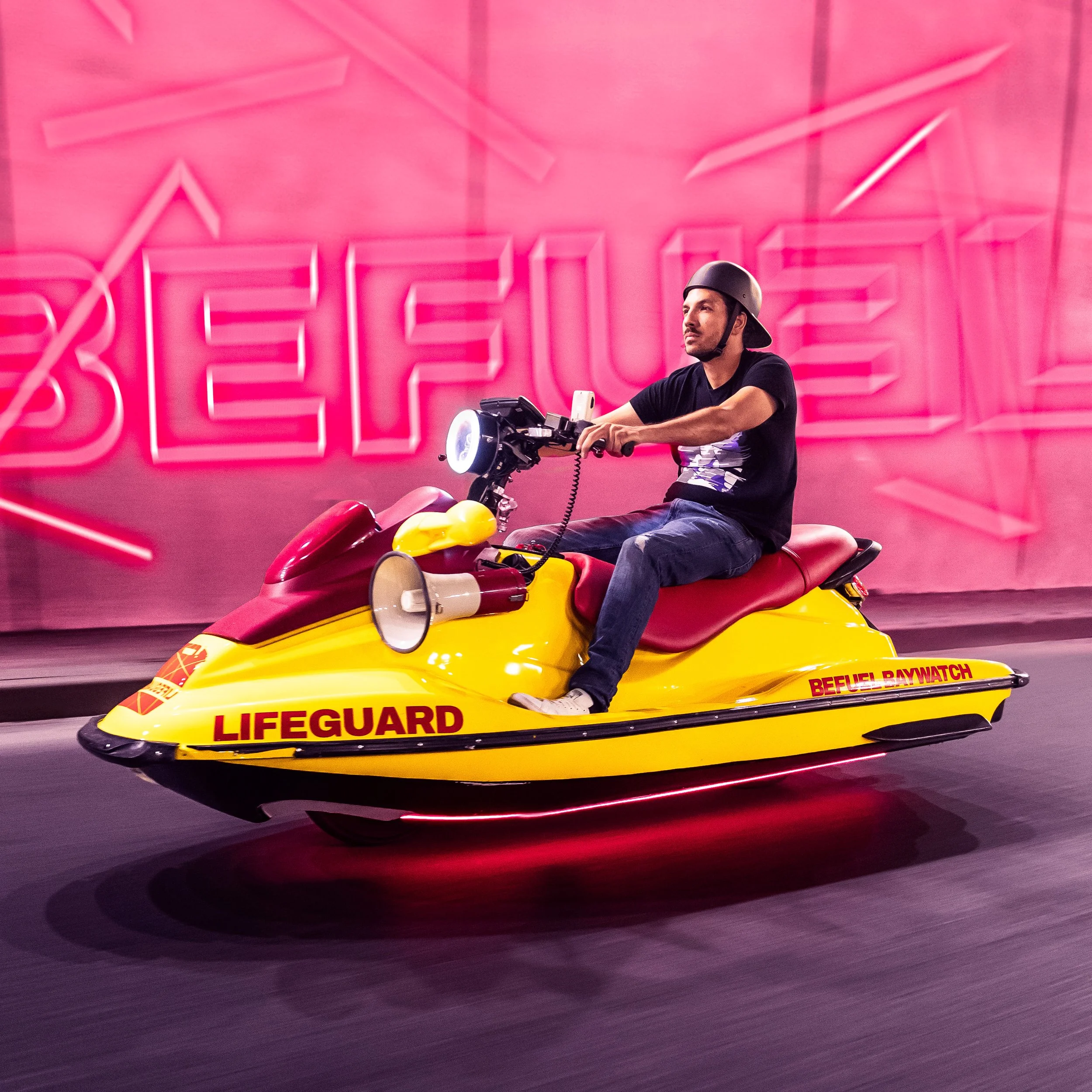 Uomo che guida un motoscooter giallo con scritte 'LIFEGUARD' e 'BEFUEL BYWATCH', indossa un casco nero e un T-shirt nero, attraversa una strada con sfondo rosa con la parola in neon 'BEBEEL'.
