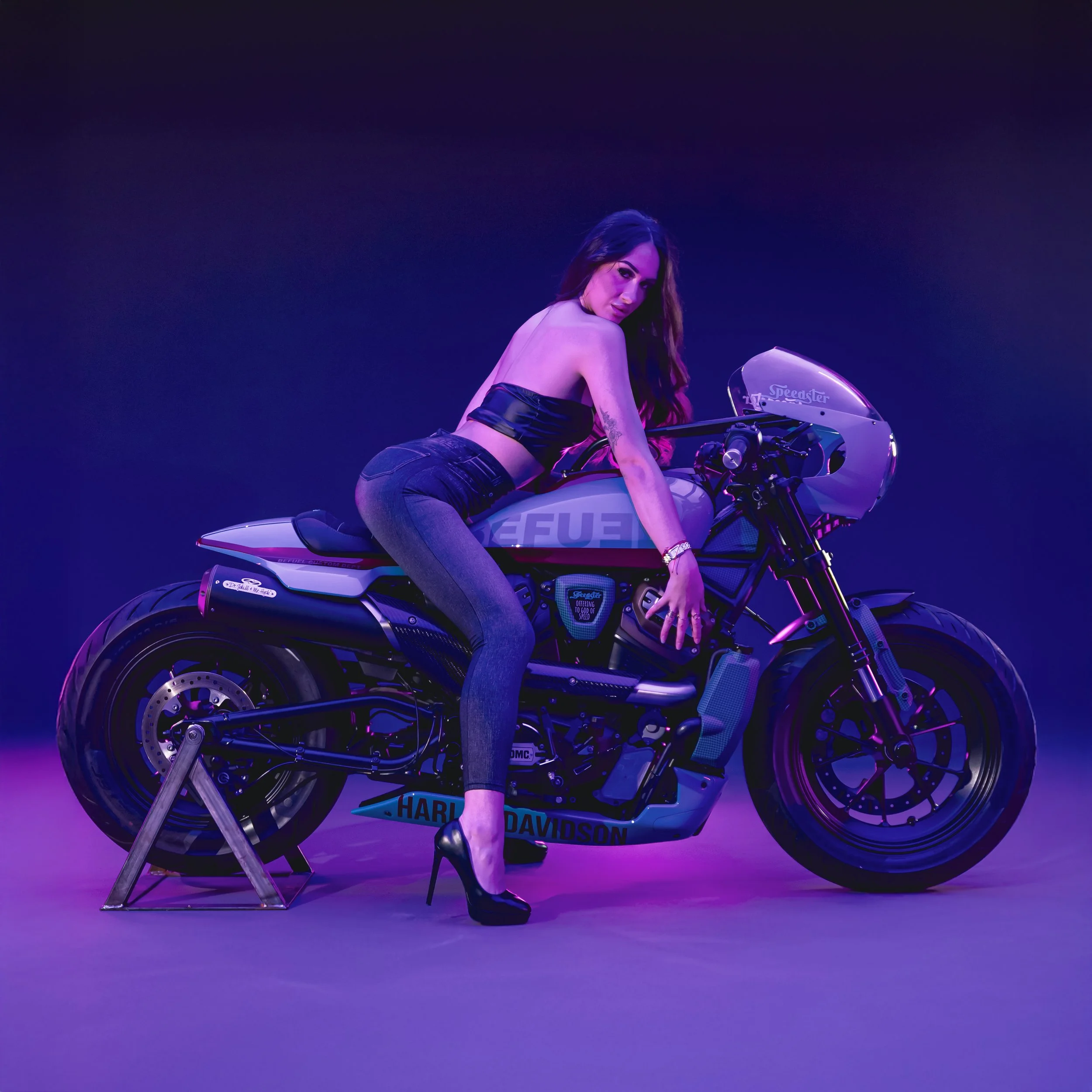 Una donna seduta su una motocicletta Harley Davidson, illuminata con luci viola e blu, sfondo scuro, indossa tacchi alti e pantaloni stretti.