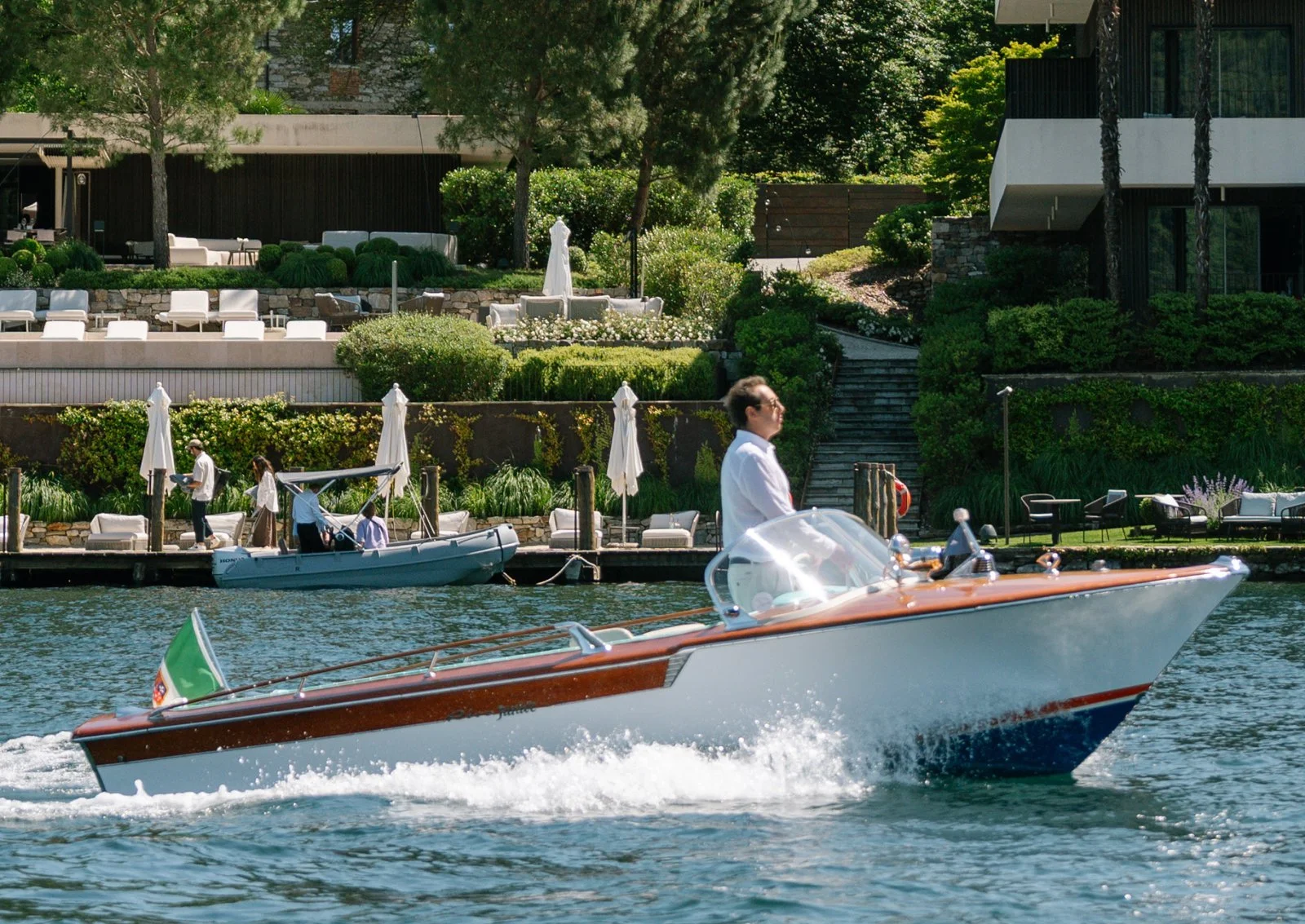 Uomo che guida una barca a motore su un lago, con case e giardini lussureggianti sullo sfondo.