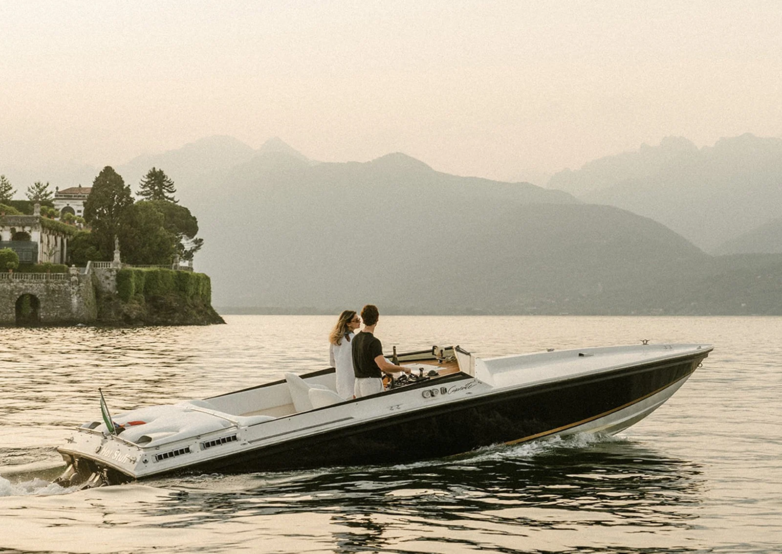 Due persone su un motoscafo navigano in un lago con montagne sullo sfondo, al tramonto.