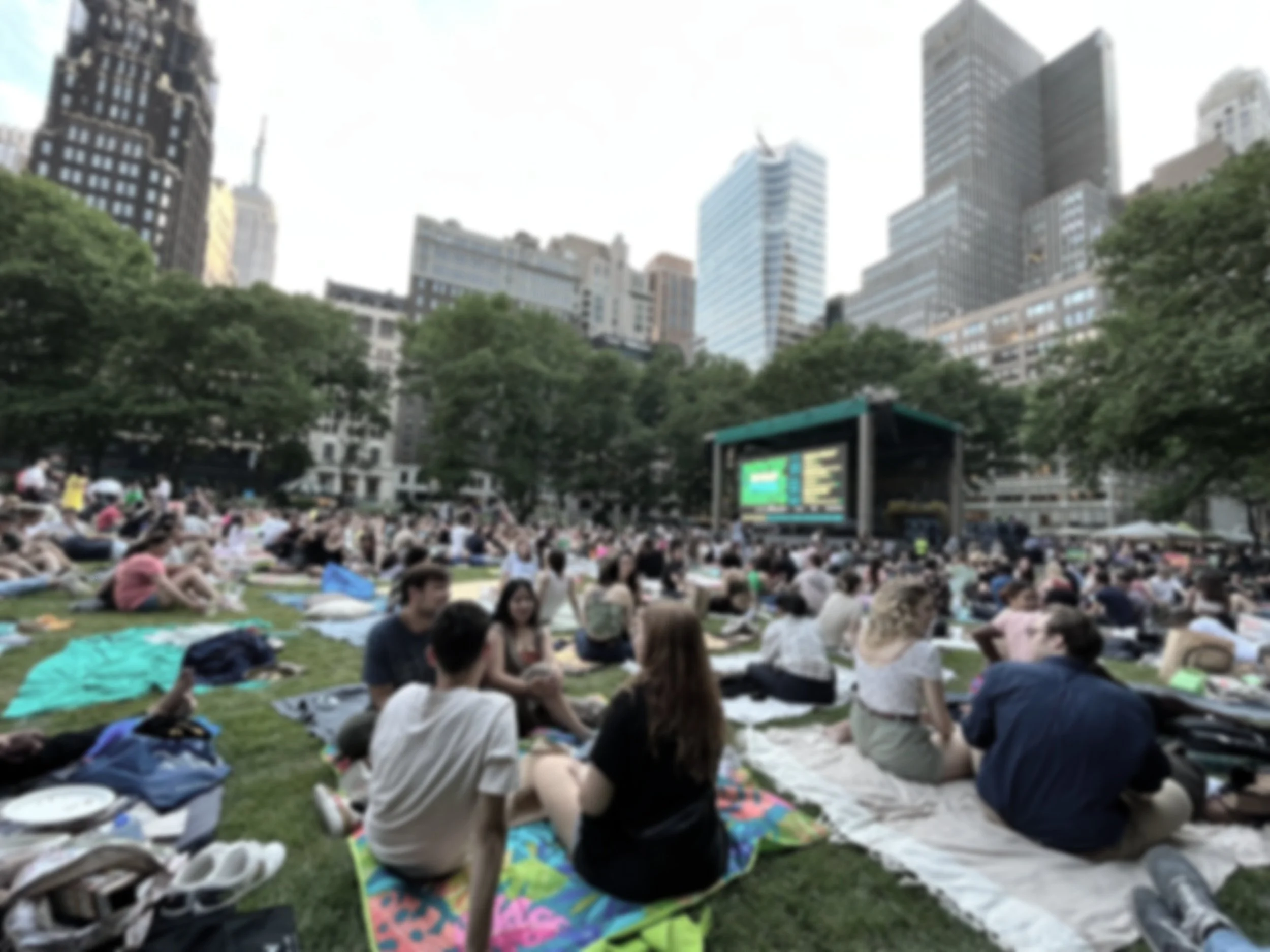 People sitting on blankets and chairs in a park, watching a large outdoor movie screen, with tall city buildings in the background.
