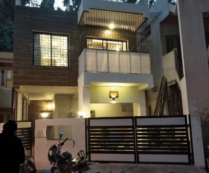 Modern multi-story house with balconies, exterior lighting, a wooden fence, motorcycles parked outside, and construction materials nearby.