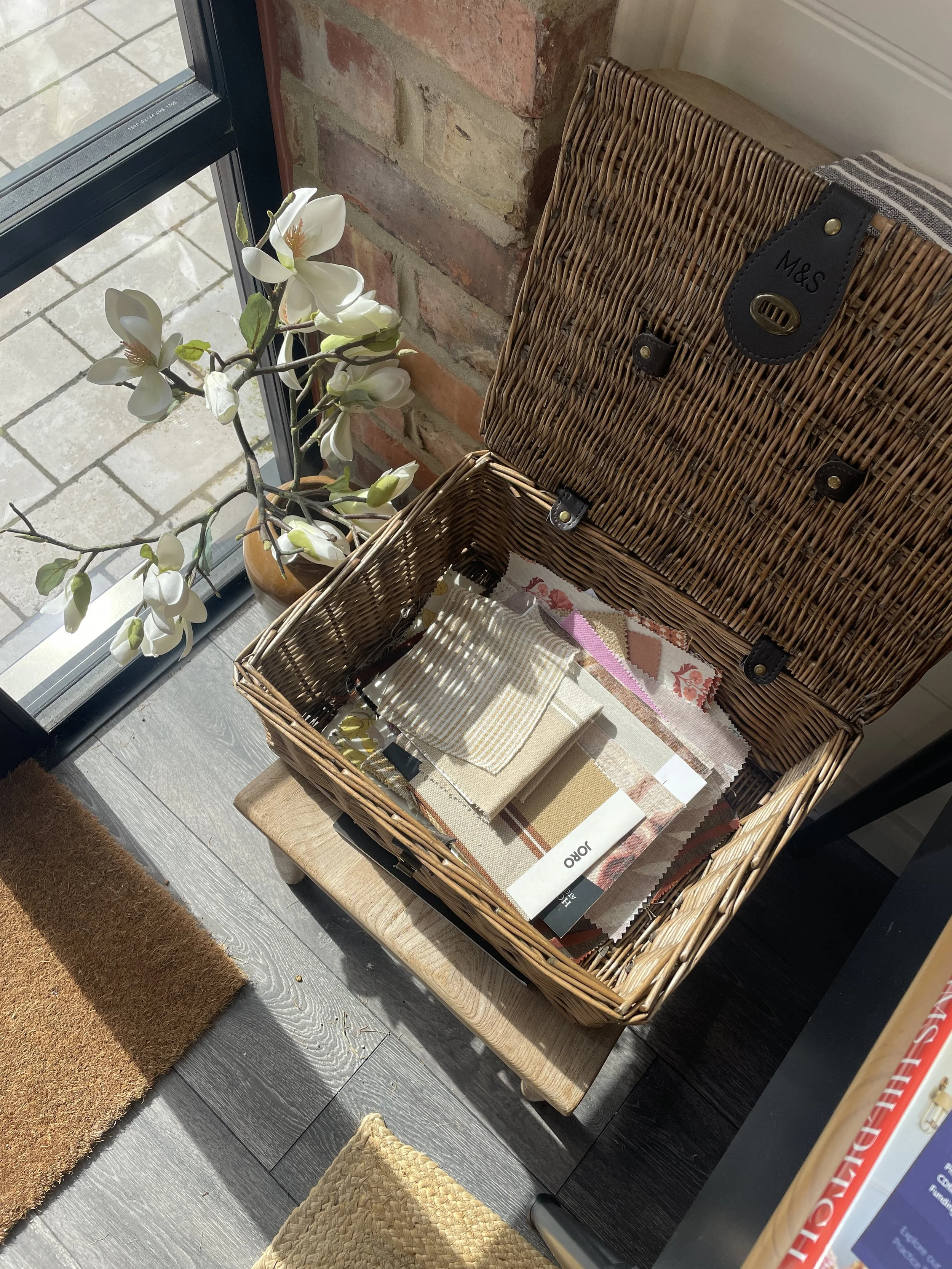 A wicker hamper of fabric samples open on a stool by a door, with magnolia stems on the windowsill behind.