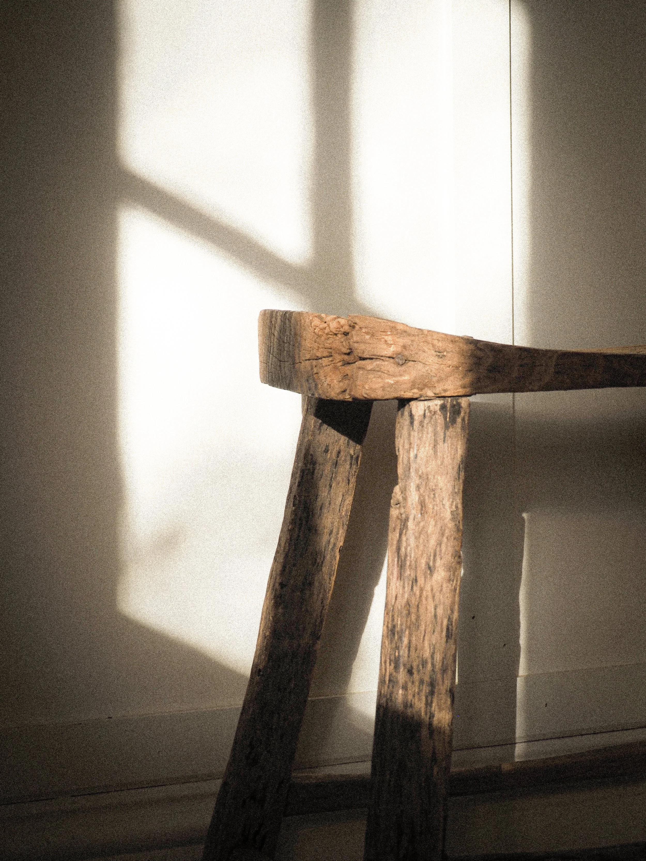 A weathered wooden stool against a pale wall in strong window light.