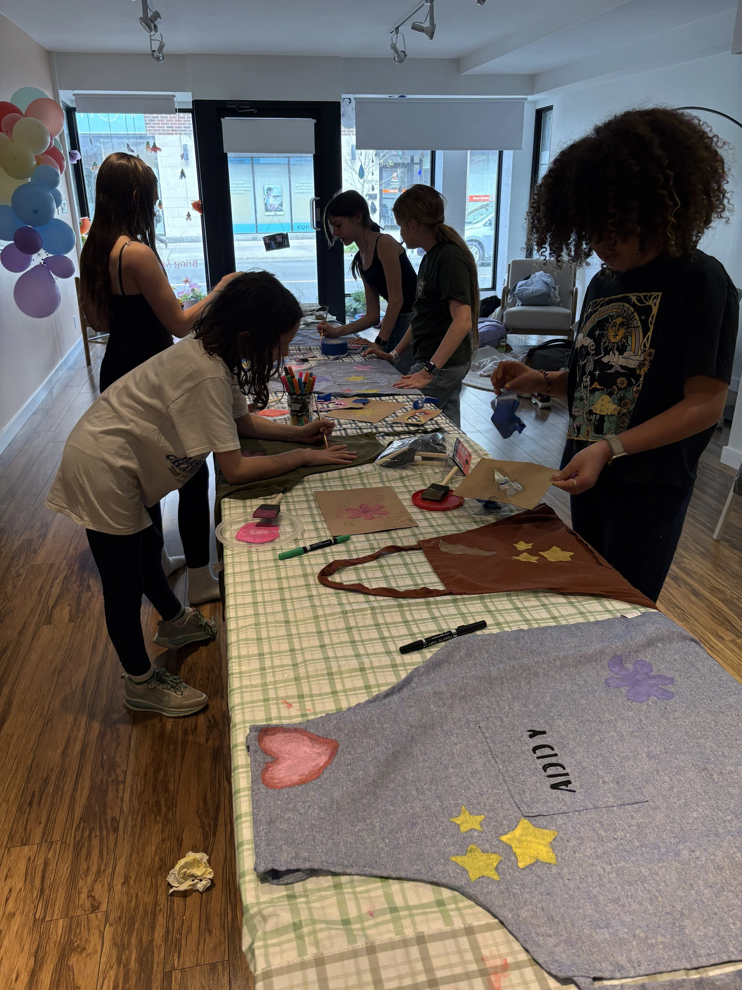 Kids working together at a long table using fabric paint to customize smocks in a bright, modern workshop space
