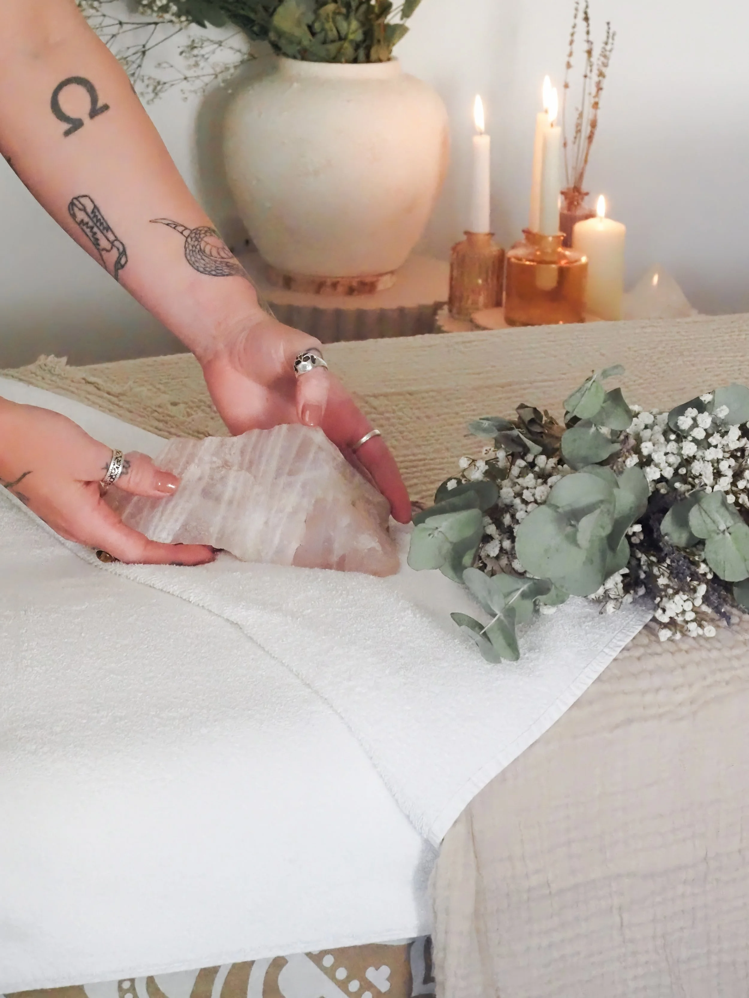 Person placing a large crystal or stone on a table decorated with flowers, with lit candles and a large ceramic vase in the background.