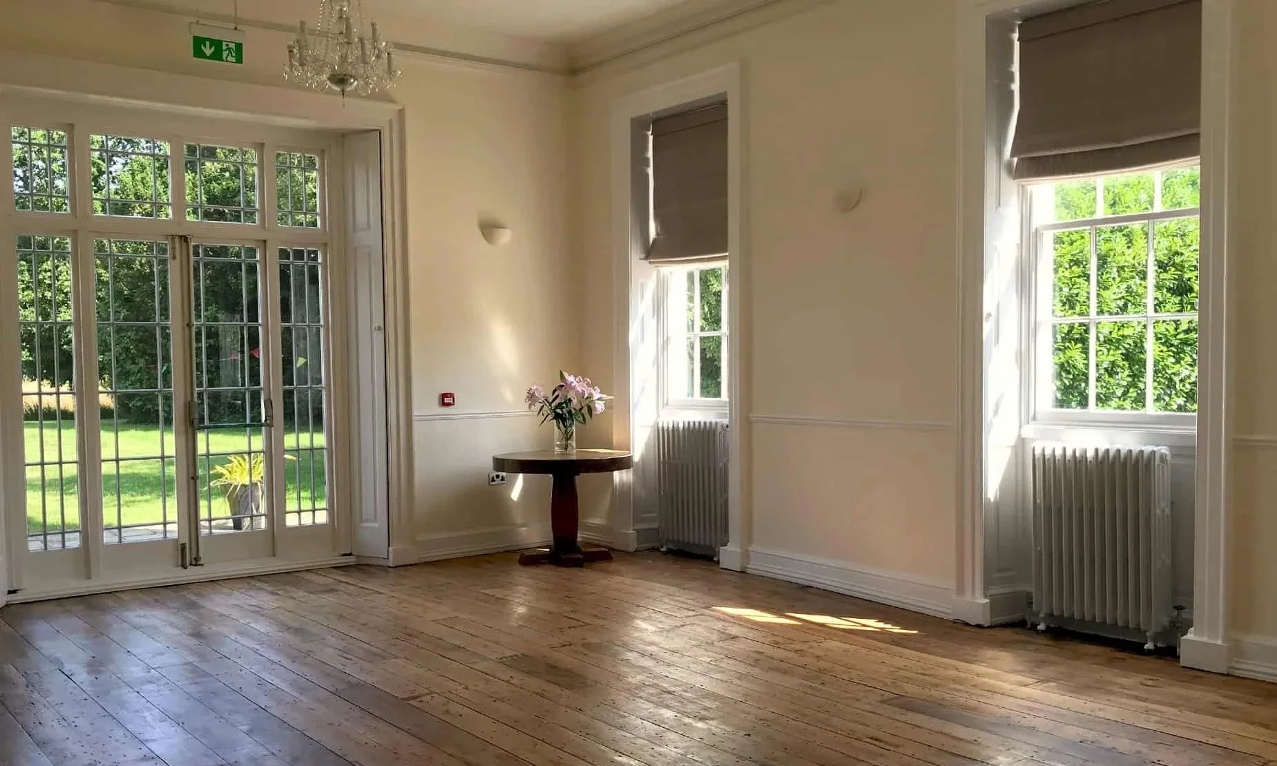 A bright empty room with wooden floors, three windows with shades, a double glass door leading outside, a chandelier in the ceiling, and a small table with a flower vase.