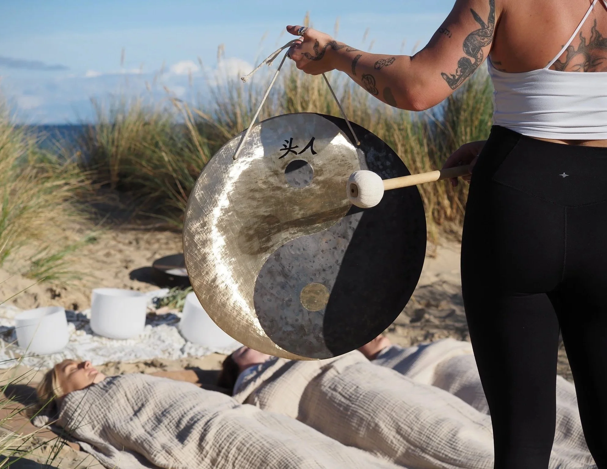 Person playing gong on beach with two people lying nearby under a blanket, ocean and sand dunes in background.