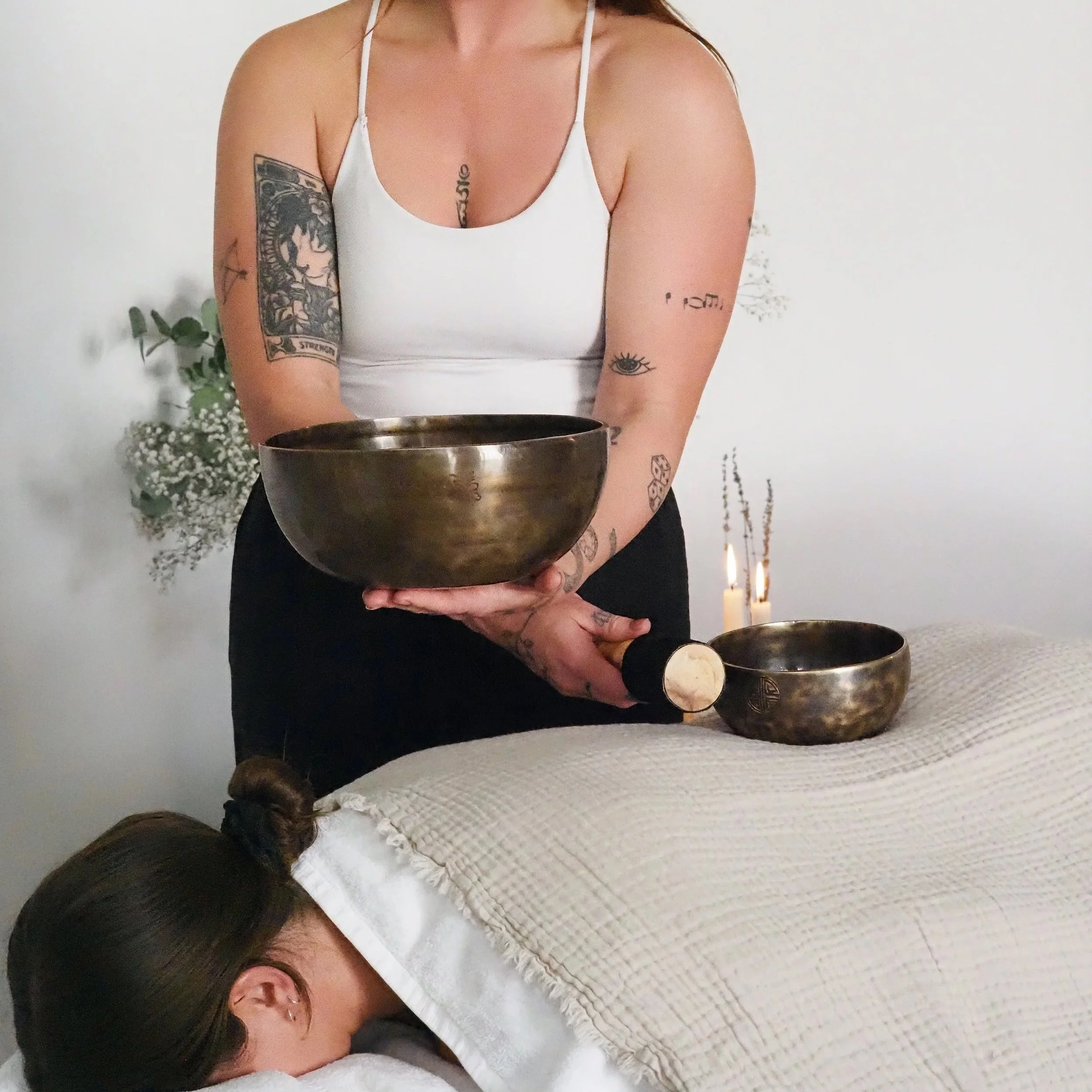 A woman performing sound therapy with Tibetan singing bowls while another person receives a massage underneath a white blanket.