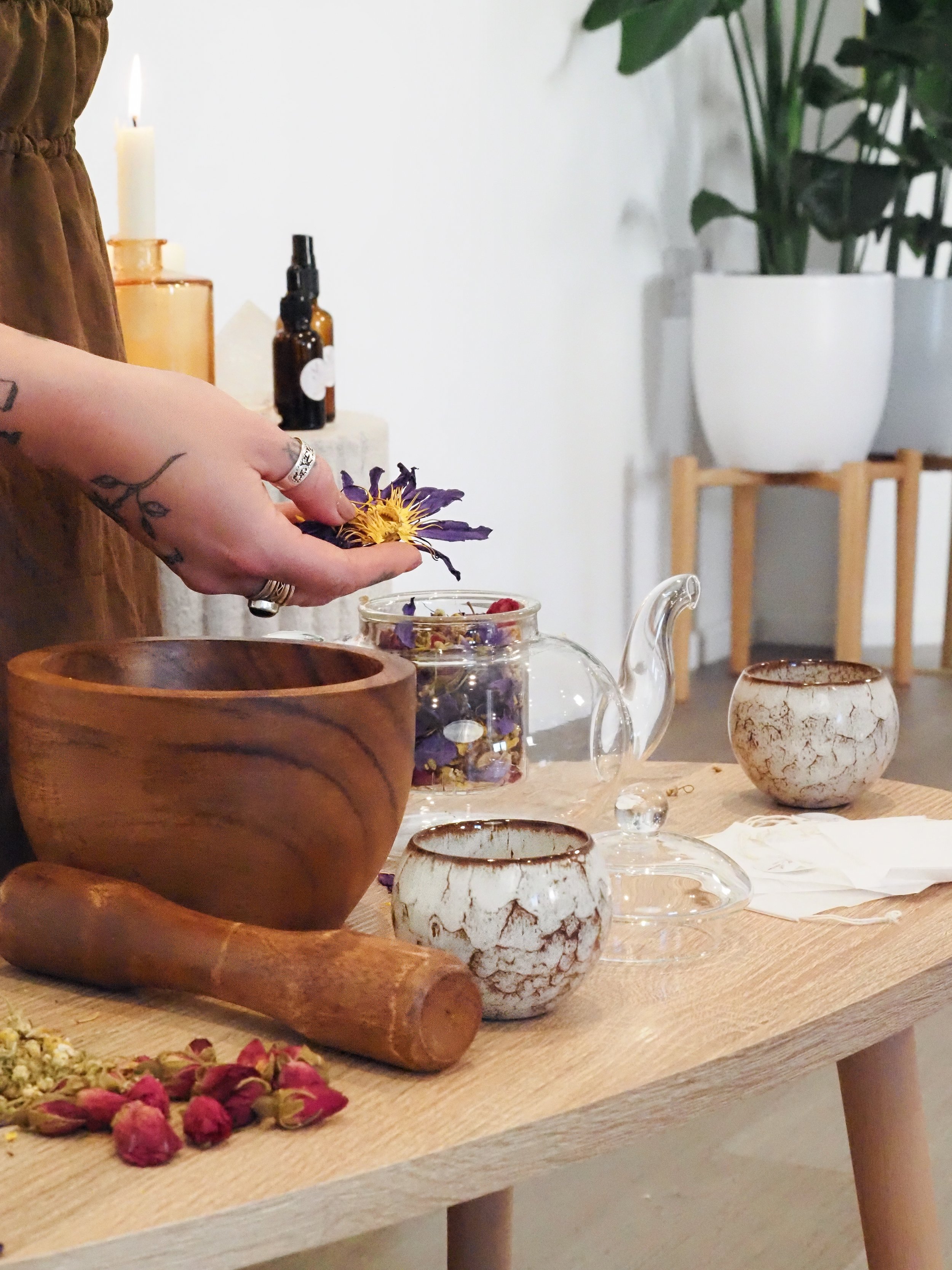 A person handling dried purple flower petals over a wooden table with ceramic bowls, a glass teapot, a glass jar filled with dried flower petals, and a wooden pestle. The background shows potted plants and decorative bottles.