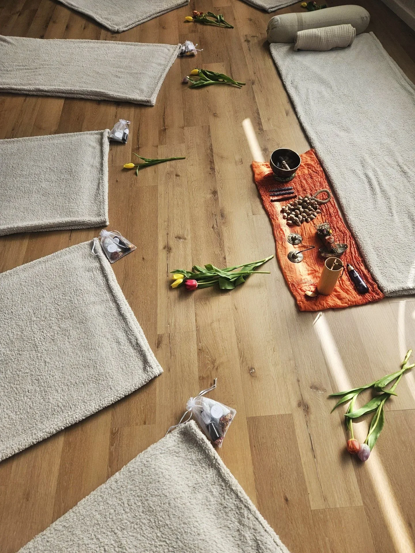 A beginner's yoga or meditation setup with blankets on a wooden floor, tulip flowers, a red cloth with various items like stones, candles, and incense, and some small gift bags.