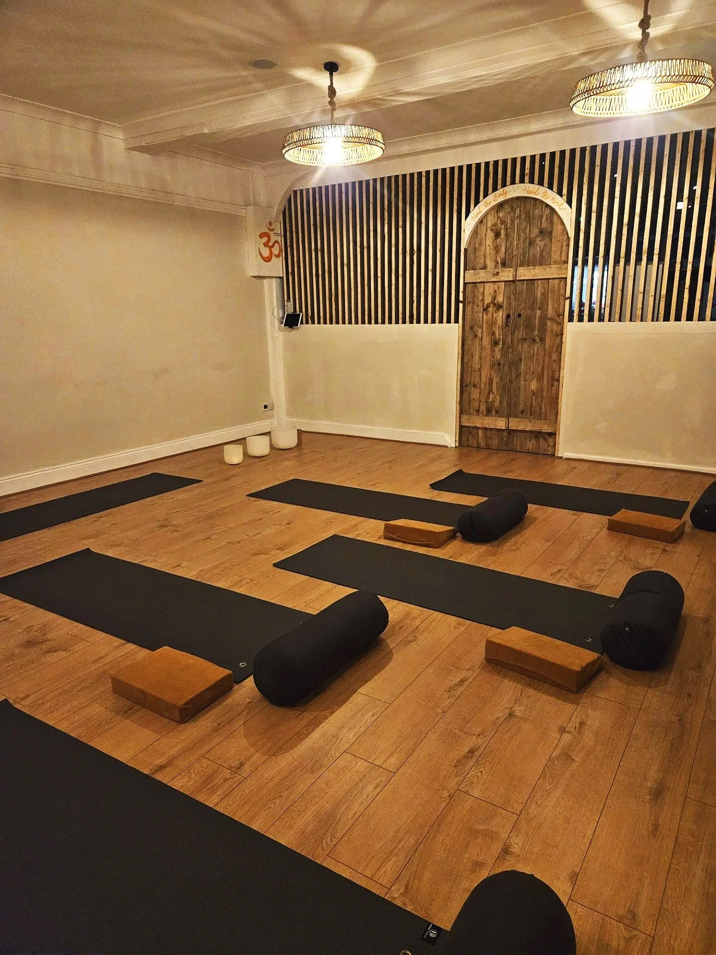 Yoga class setup with black mats, bolsters, blocks, and cushions on a wooden floor in a dimly lit room with hanging pendant lights and a wooden door at the back.