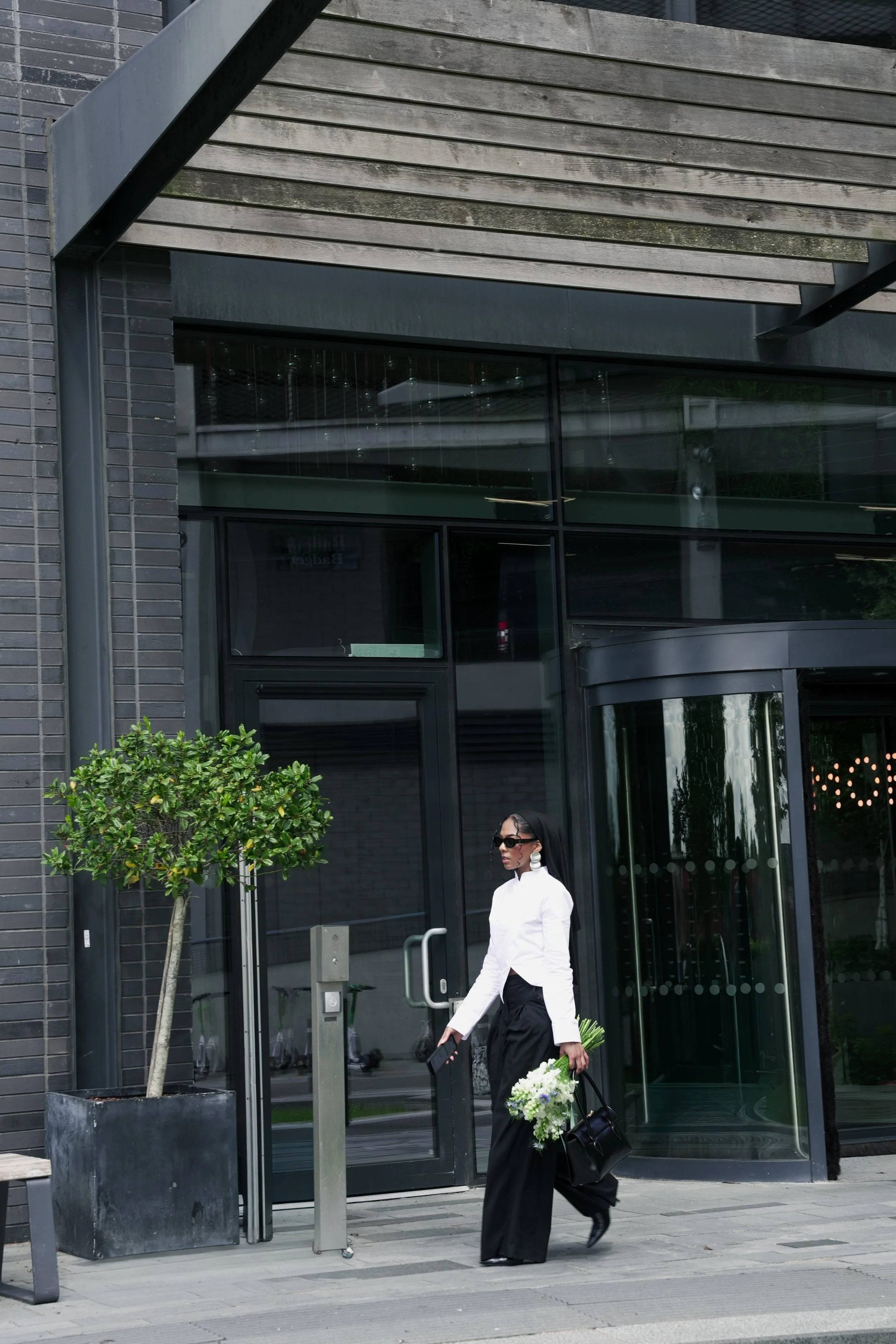 Woman holding a bouquet of flowers and a bag, walking past a modern glass building entrance in an urban setting.