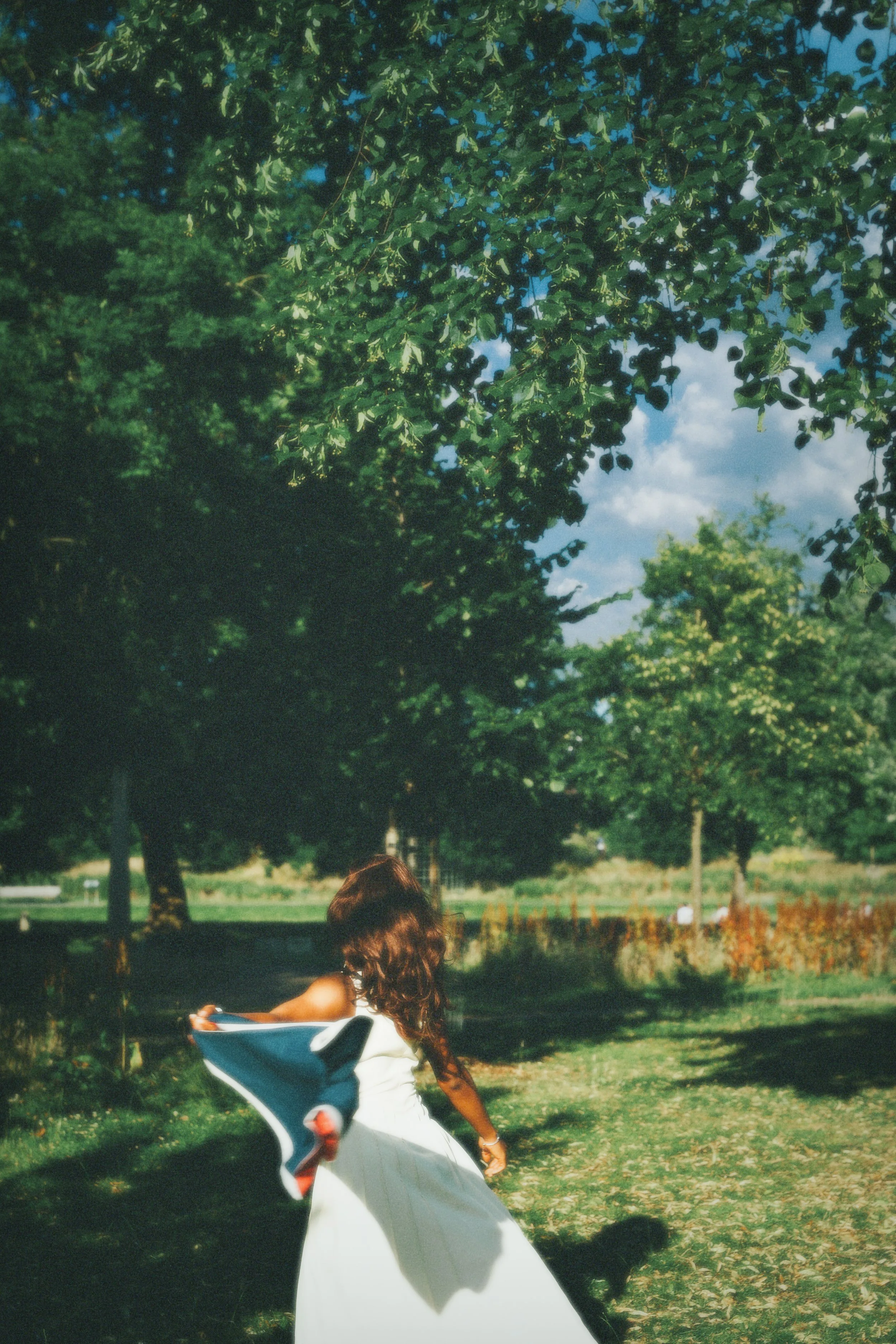 A woman with long brown hair in a white dress is sitting outdoors in a park, surrounded by trees and green grass, with her back to the camera.