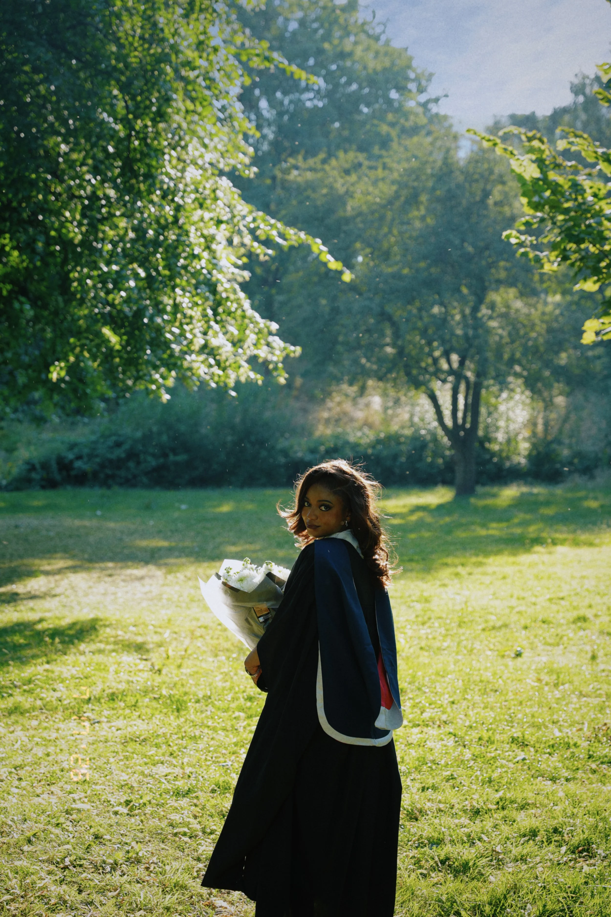 A woman in a graduation gown holding a bouquet of flowers outdoors on a sunny day with green trees and grass in the background.