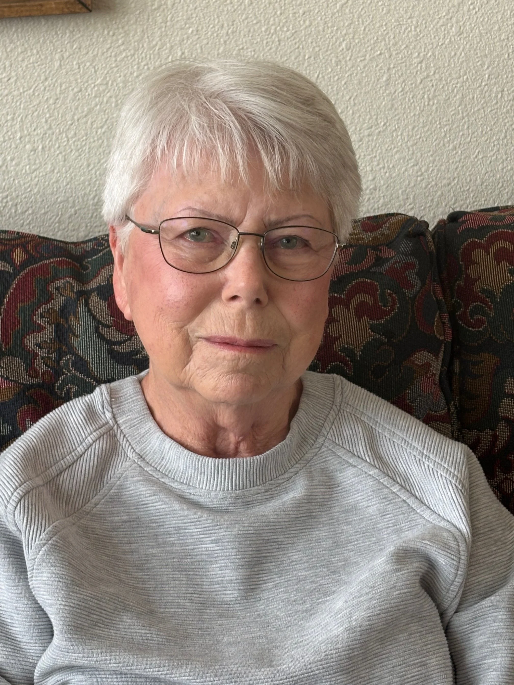 A woman with glasses and short white hair, wearing a red top, standing indoors.