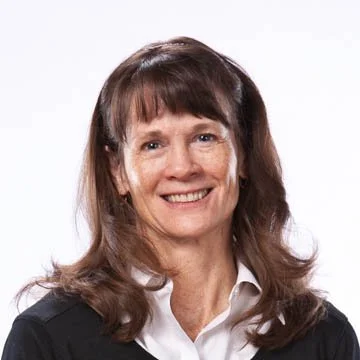A woman with brown hair and bangs smiling, wearing a white collared shirt and black sweater against a plain white background.