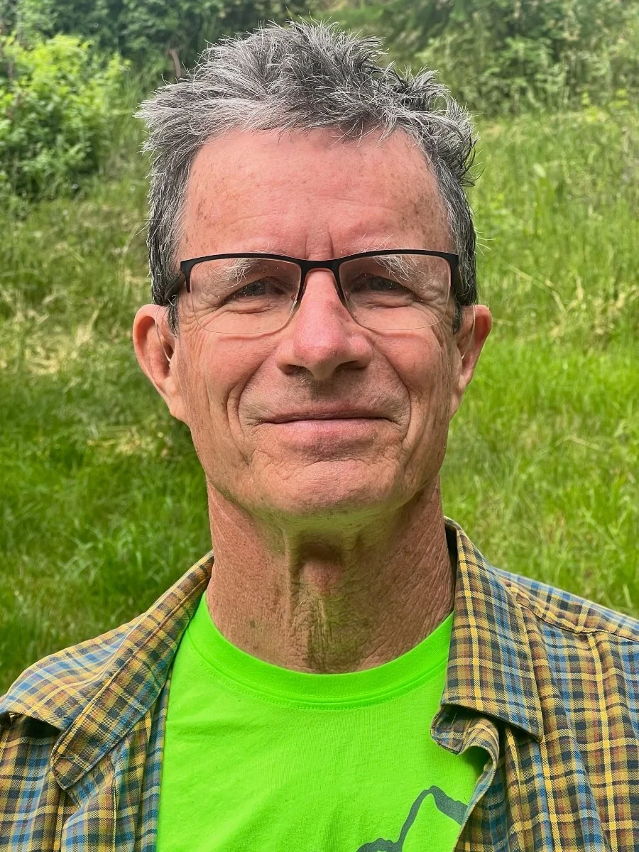 A middle-aged man with glasses, a beard, and short hair, smiling against a green background.