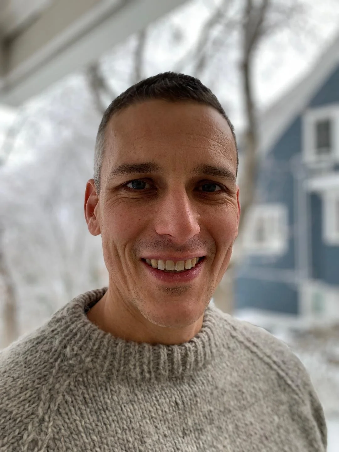 Close-up of a smiling man with short dark hair wearing a beige sweater, with blurred outdoor winter background including snow and trees.