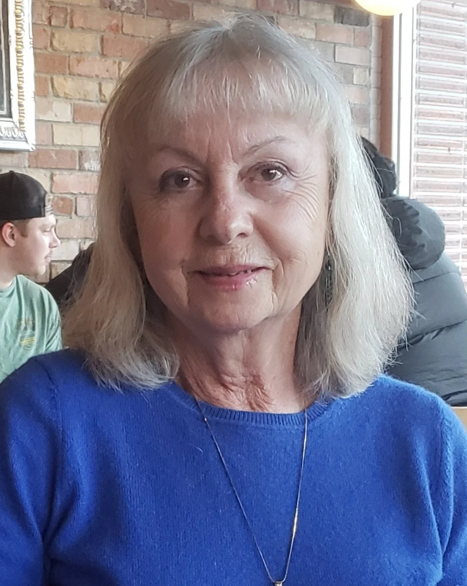 An elderly woman with shoulder-length blonde hair wearing a blue sweater and a necklace, sitting inside a restaurant with brick walls and large windows.