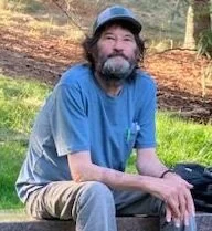 Portrait of a man with curly dark hair and a mustache, wearing an orange jacket and white shirt, smiling