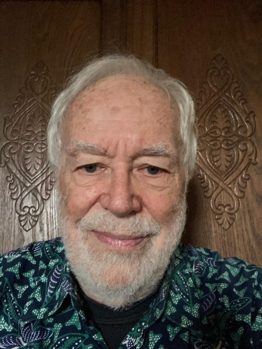 Older man with white hair and beard smiling in front of carved wooden background.
