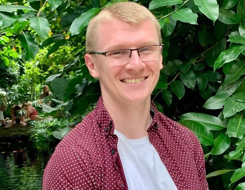 A smiling young man with glasses and blonde hair, wearing a white t-shirt and a maroon shirt with white polka dots, standing in front of lush green tropical plants.
