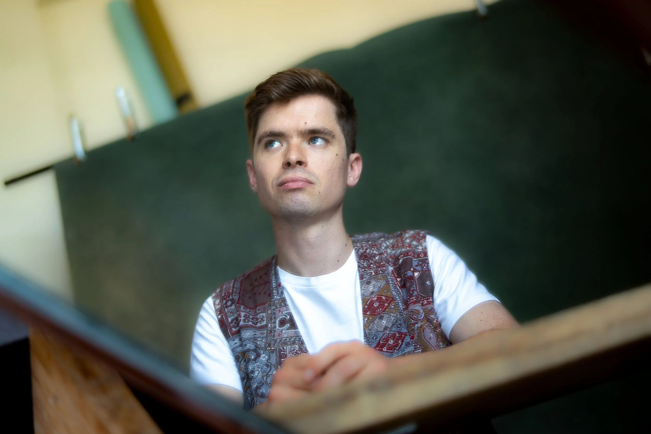 A young man with brown hair and blue eyes is sitting at a desk in front of a laptop, looking slightly upwards with a thoughtful expression.