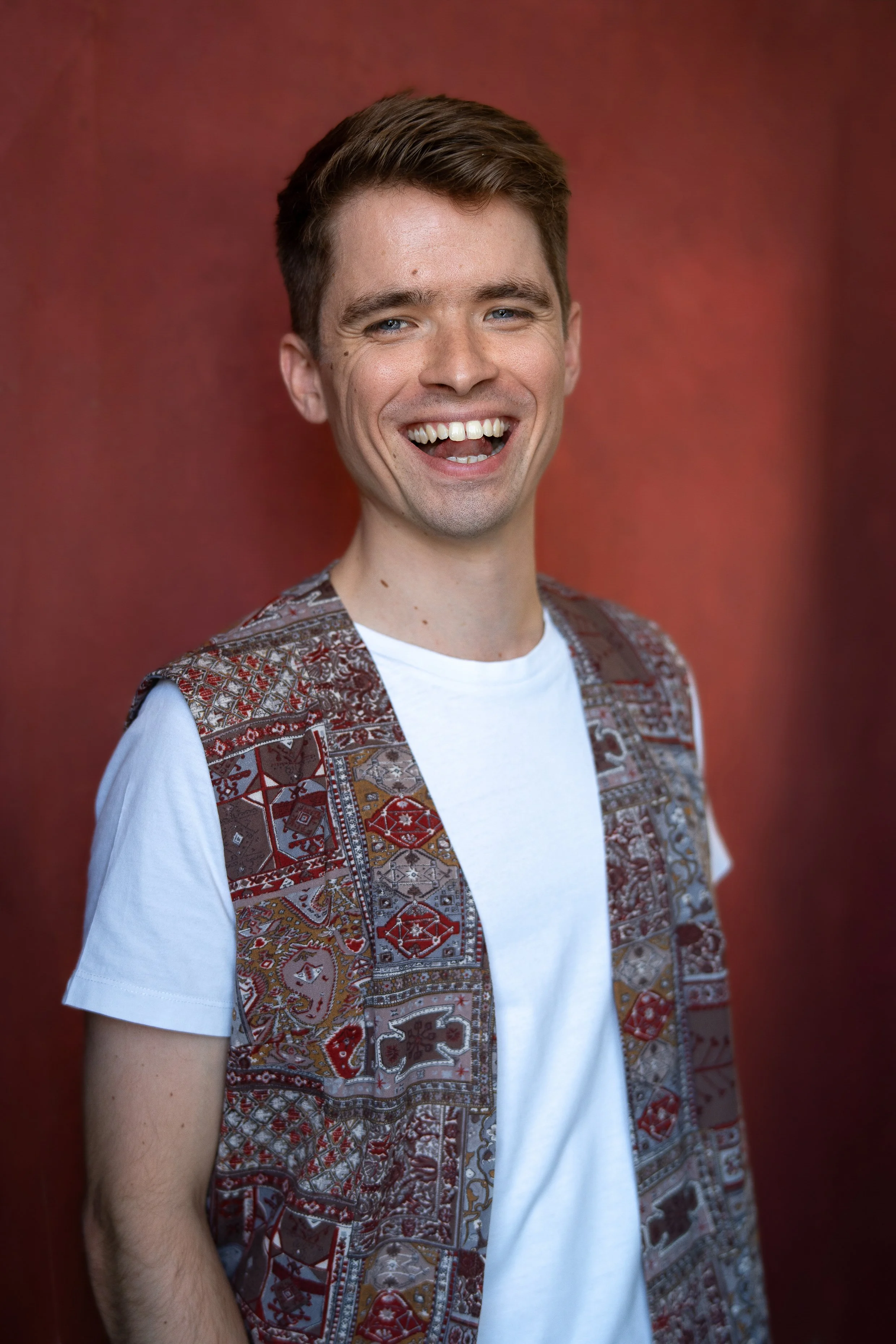 A young man with short brown hair, blue eyes, and a wide smile showing his teeth, standing against a red background. He is wearing a white t-shirt and a patterned vest with red, brown, and beige designs.
