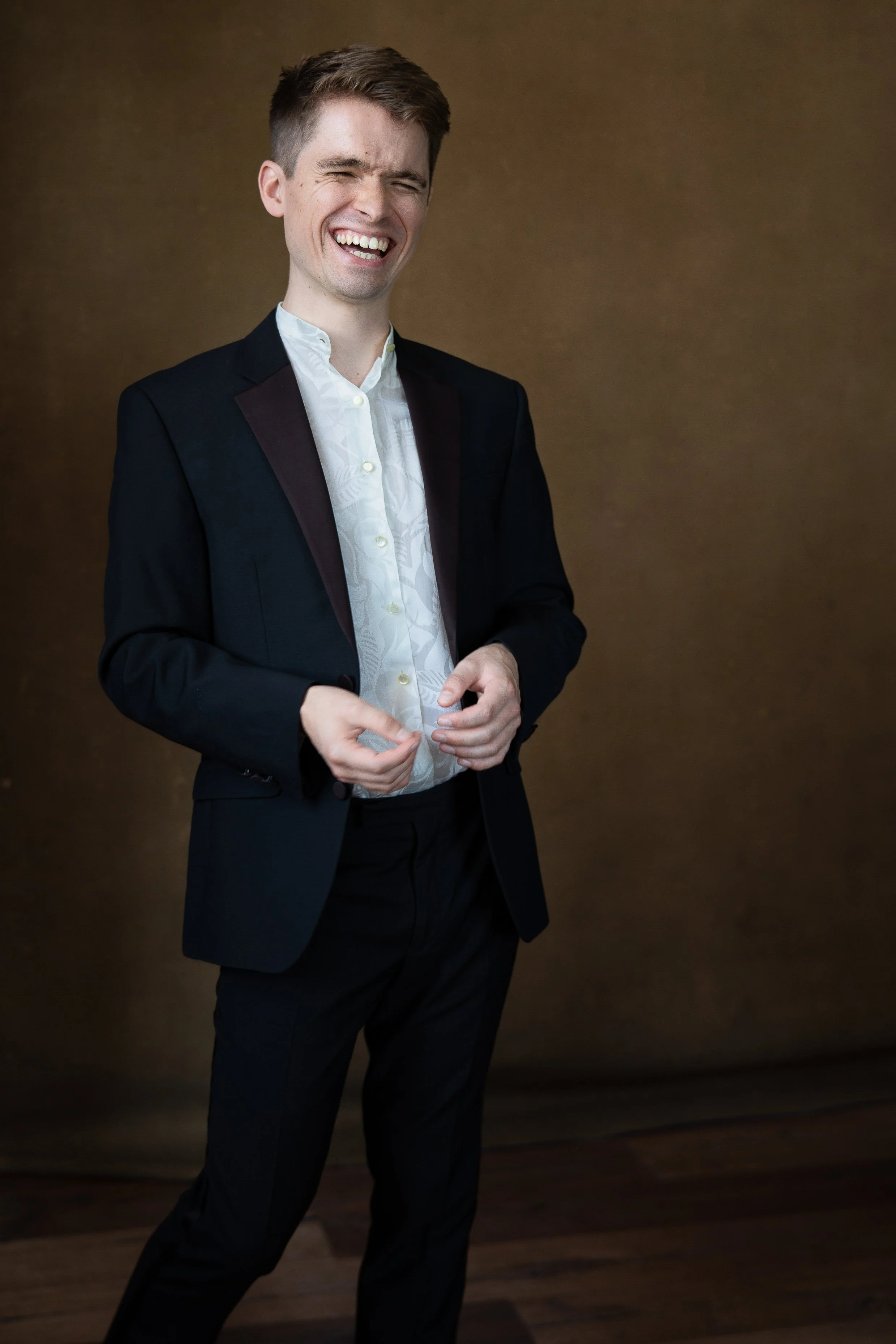 Young man in a black suit and white shirt, smiling and laughing against a brown background.