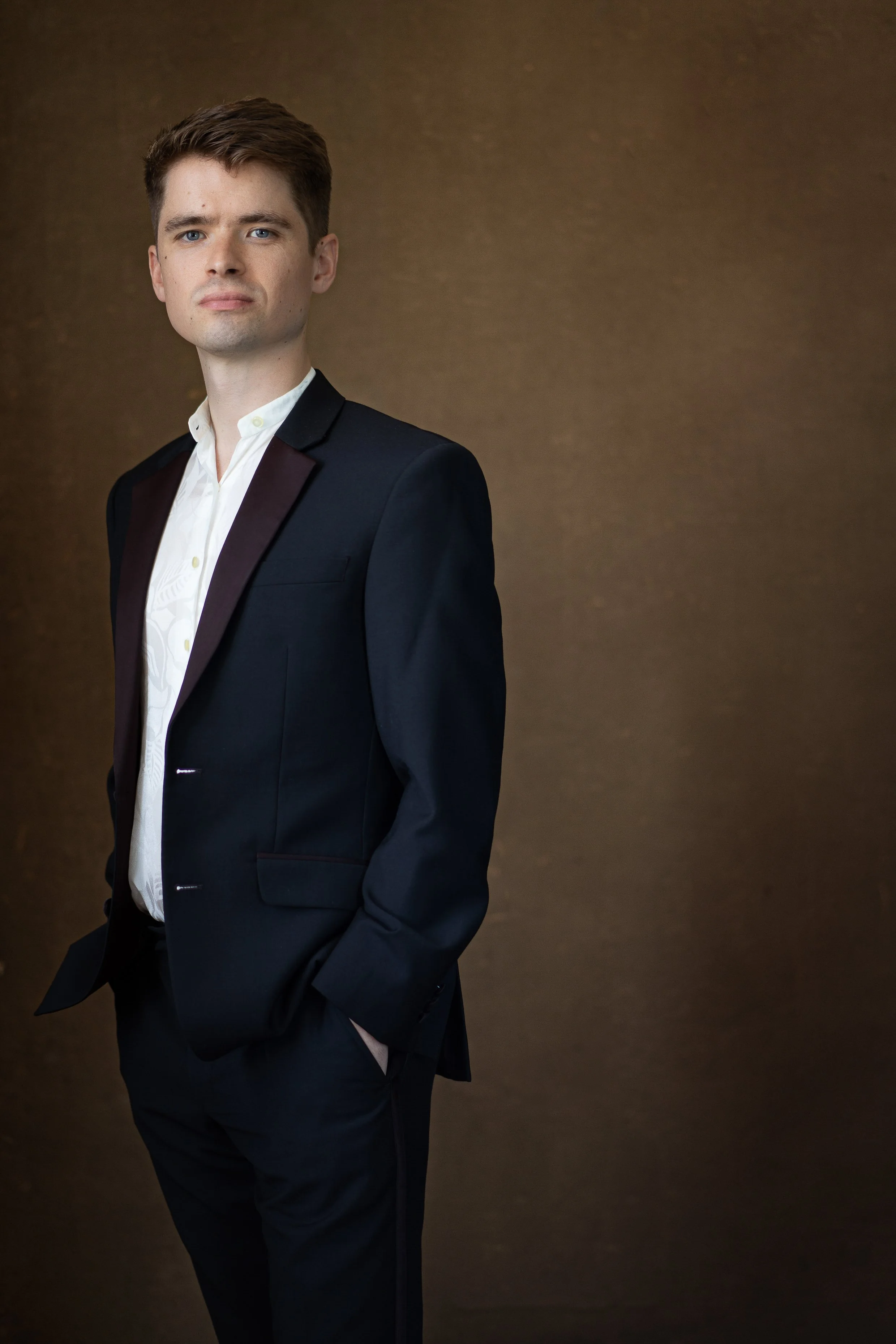 A young man in a suit standing against a brown background.