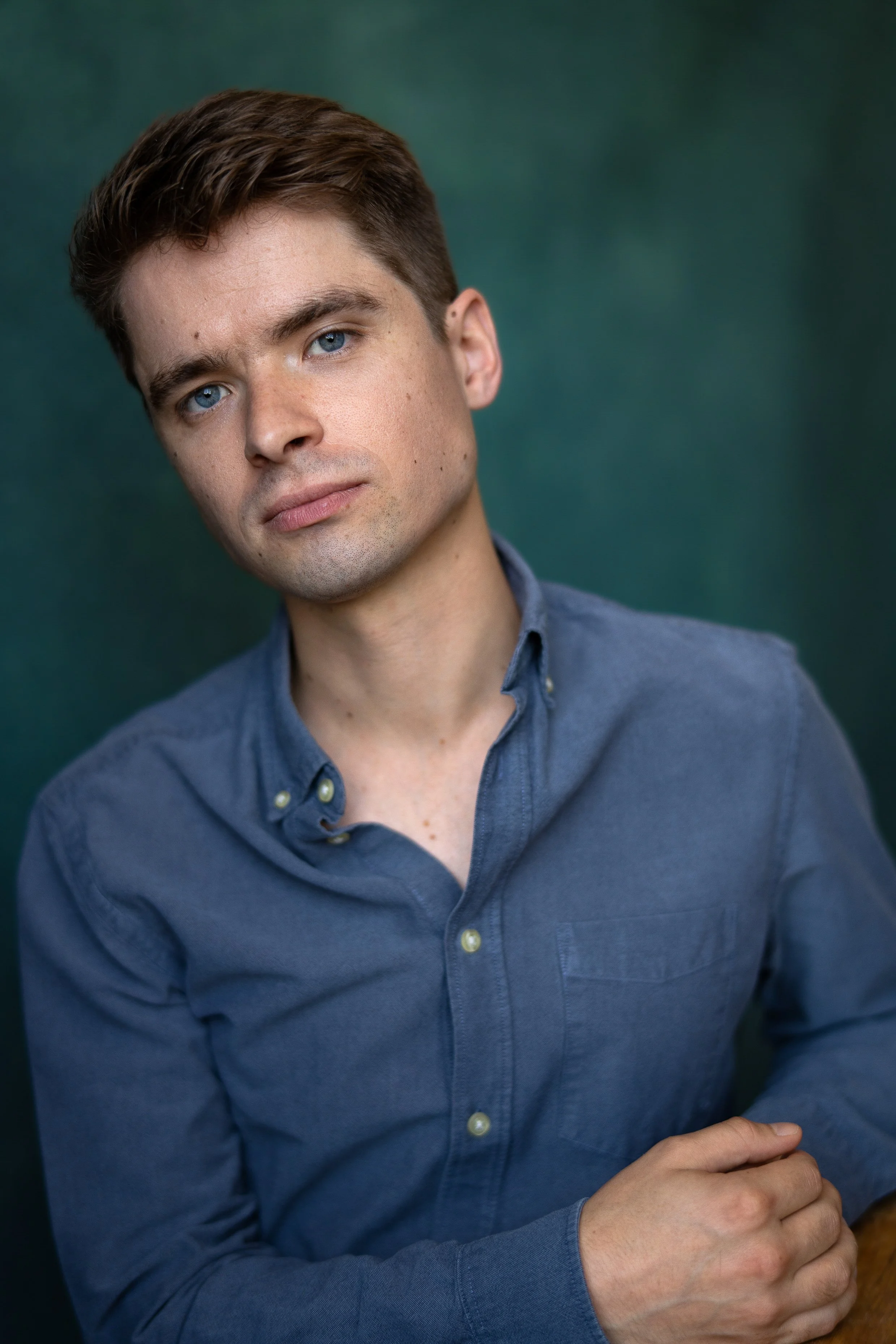 Portrait of a young man with brown hair and blue eyes, wearing a blue button-up shirt, against a dark green background.