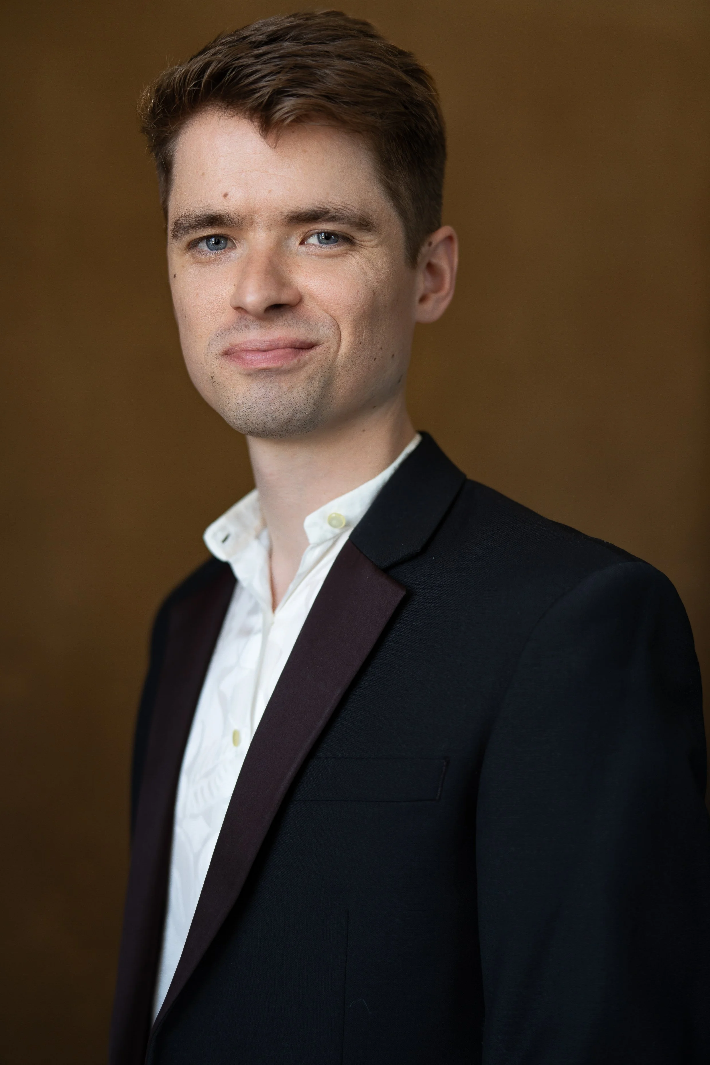 A professional headshot of a young man with light skin, blue eyes, short brown hair, wearing a dark blazer and white shirt, standing against a plain brown background.