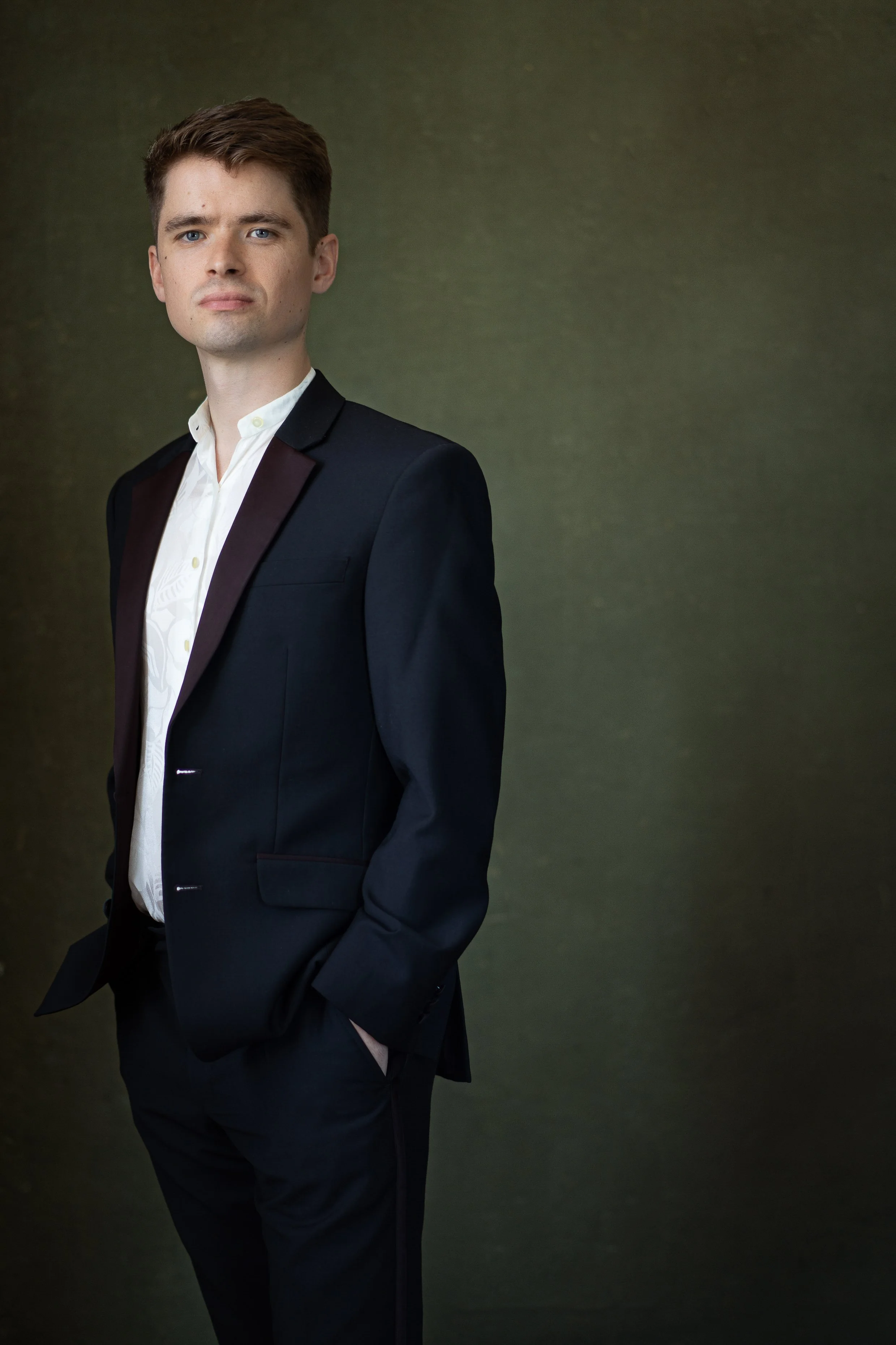 A man in a black tuxedo with a white shirt standing against a dark green background.