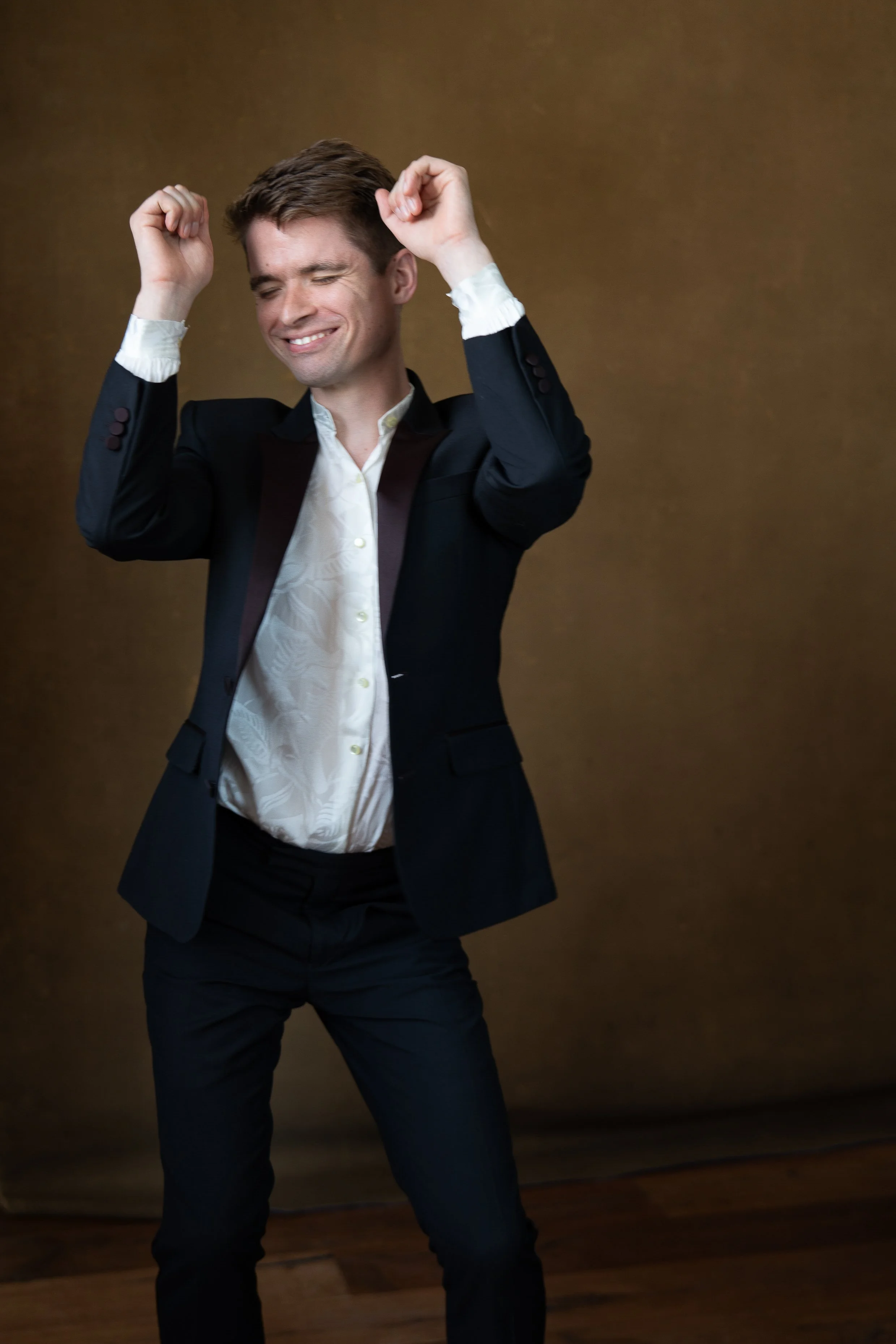 Man in a black suit with a cream-colored shirt dancing with a smile against a brown background.