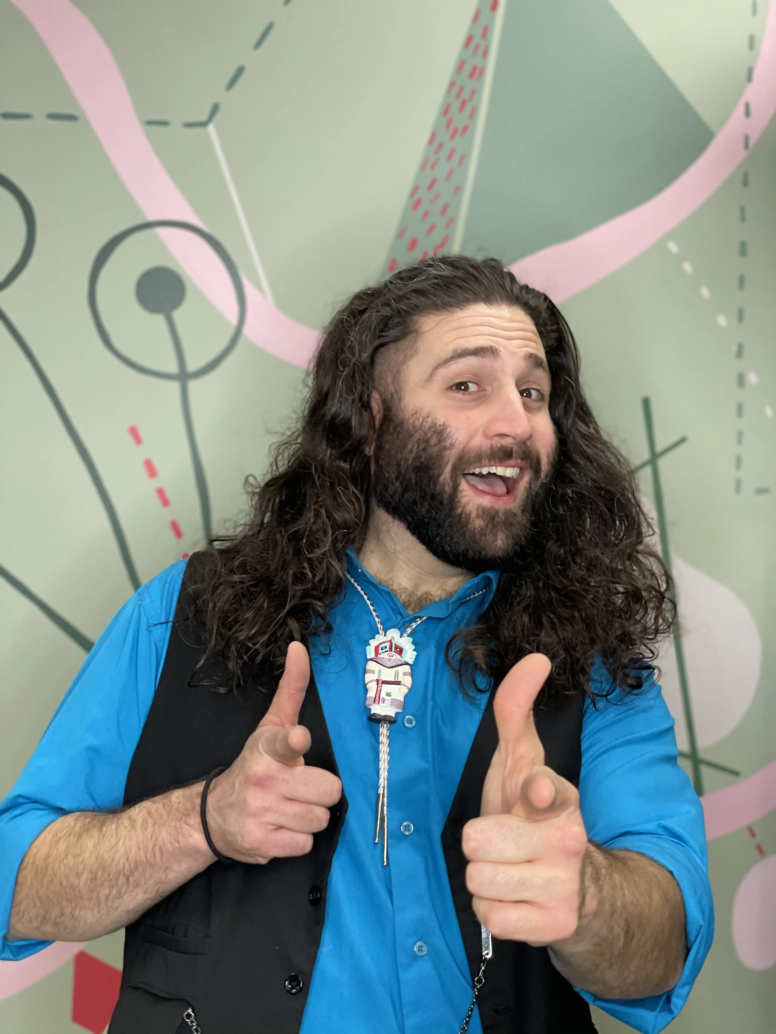 A smiling man with long curly hair and a beard giving two thumbs up, wearing a blue shirt, black vest, and a colorful Navajo totem bolotie, standing in front of colorful abstract art.