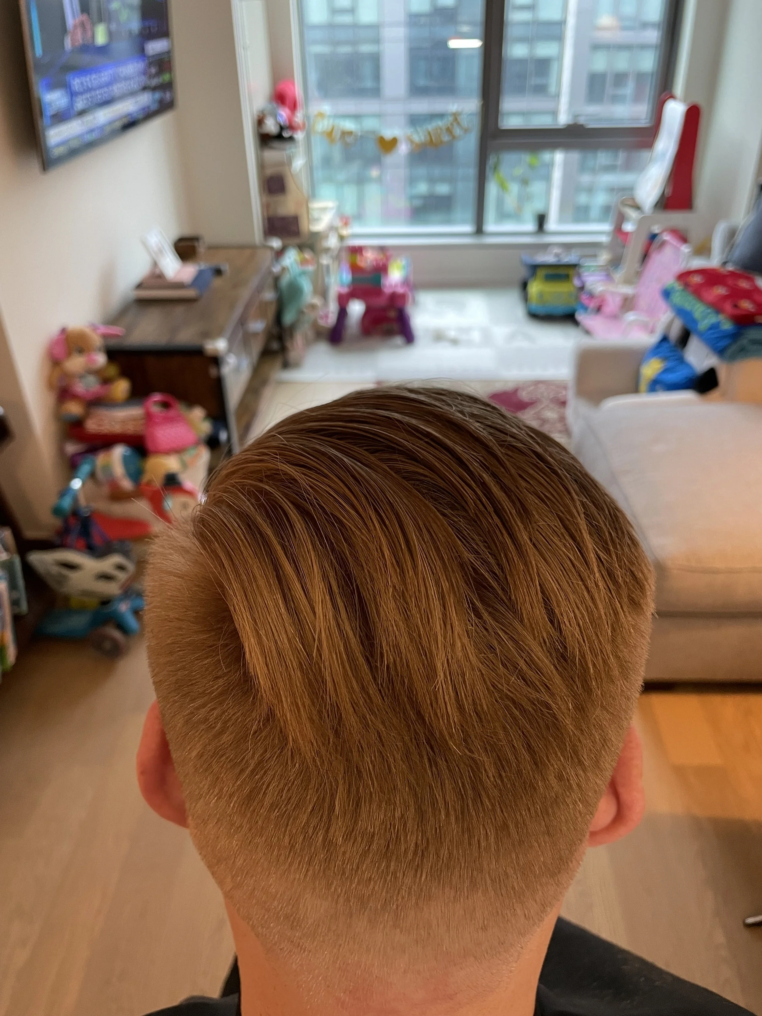 The image shows the back of a person's head with a fresh, blonde haircut, in a living room filled with toys and colorful children's items, with large windows in the background.