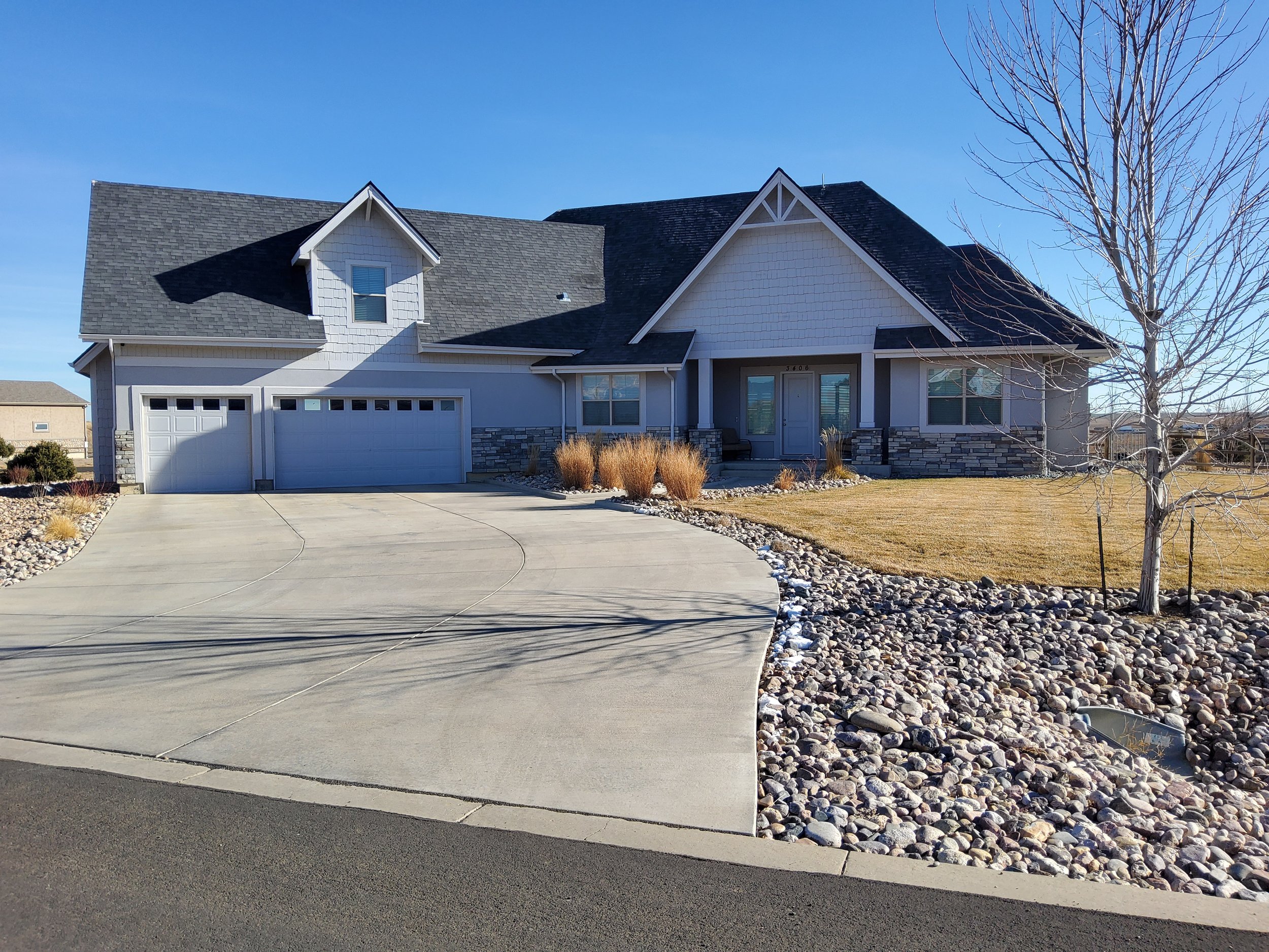 A modern suburban house with a gray roof, white siding, stone accents, and a three-car garage. The front yard has a curved concrete driveway, a leafless tree, and dry grass surrounded by rocks.