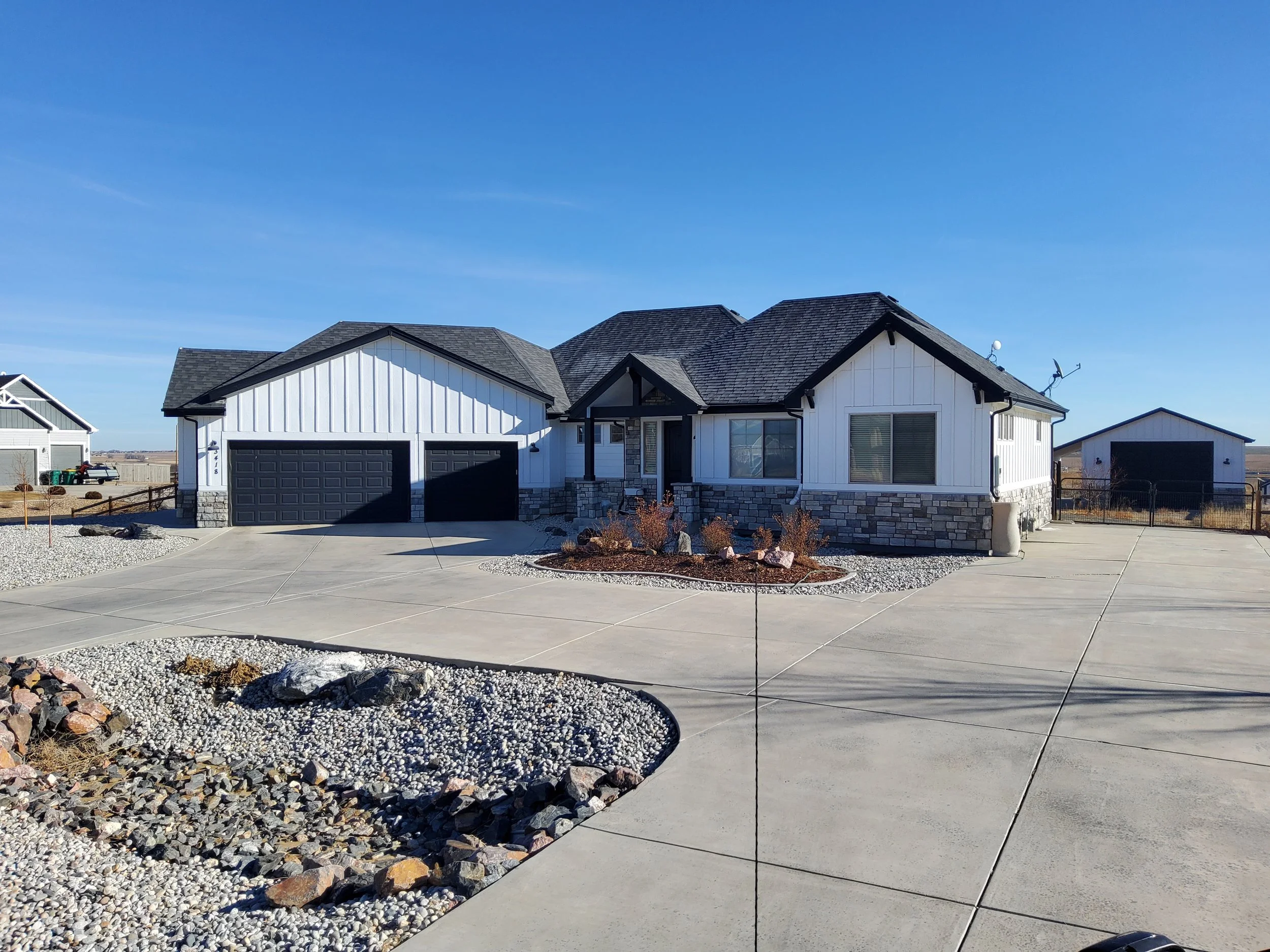 Front view of a modern single-story house with a stone and white siding exterior, black garage doors, and a landscaped front yard with rocks and gravel. Clear blue sky.