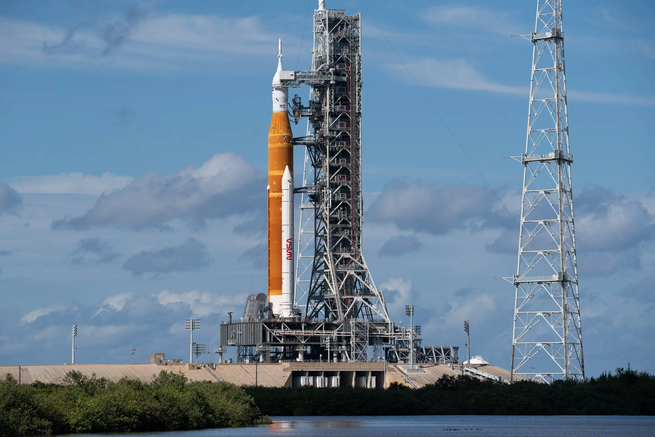 A NASA rocket on its launch pad, ready for launch, with large support structures, towers, and a cloudy sky in the background.