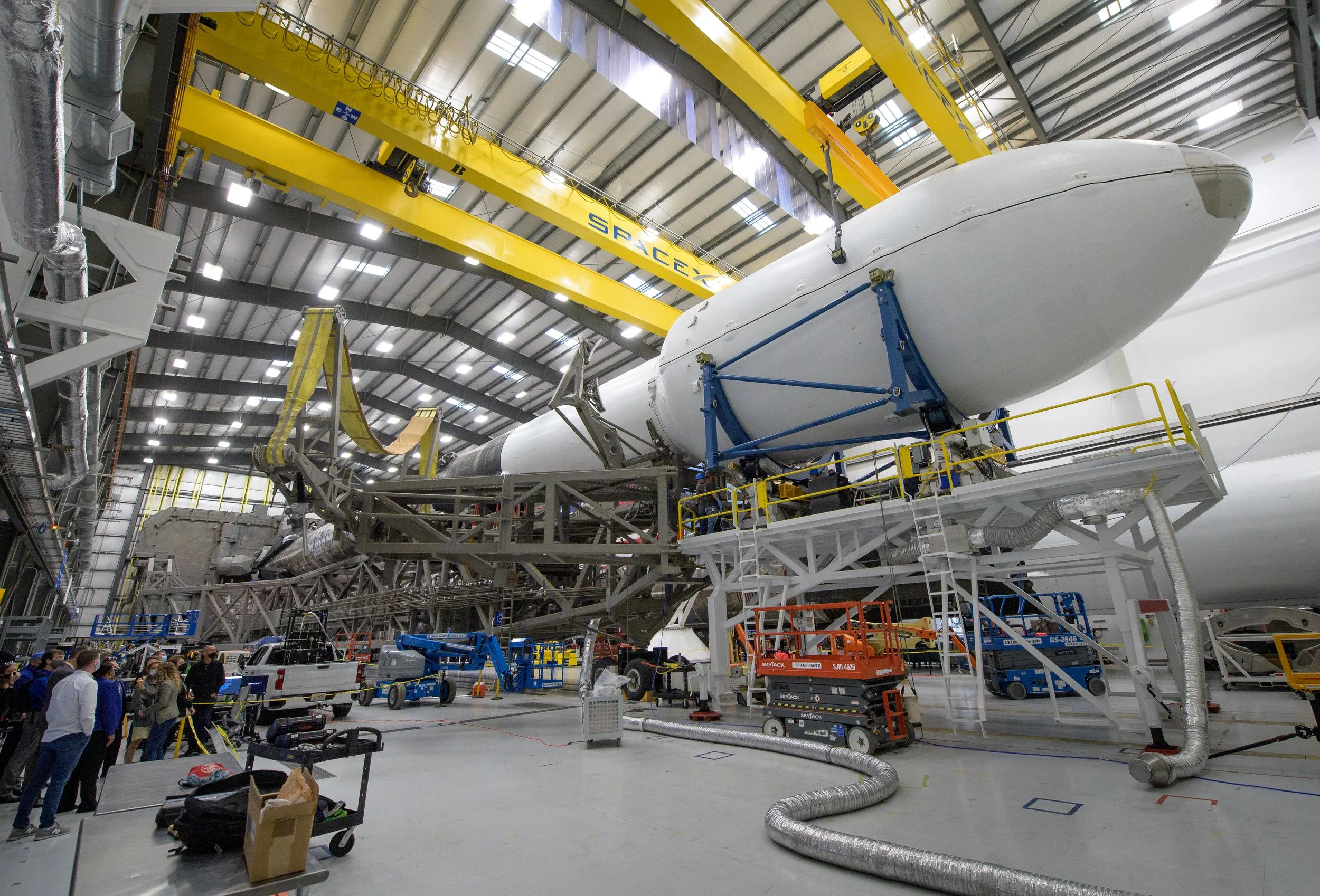 Large spacecraft or rocket being assembled inside a spacious industrial hangar. Several workers and equipment are present around the structure, with a prominent yellow overhead crane labeled 'SpaceX' assisting in the process. JWST.